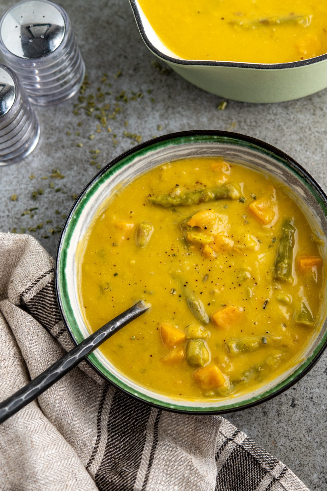 asparagus and sweet potato soup in white bowl with green, black and grey stripes, pan of soup in background