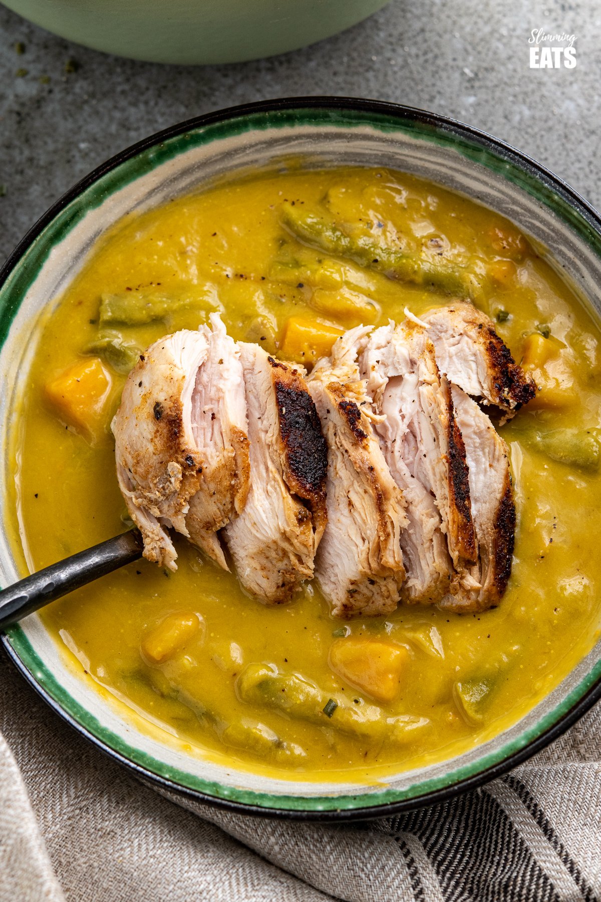 close up asparagus sweet potato soup in white bowl with green, black and grey stripes topped with sliced grilled chicken