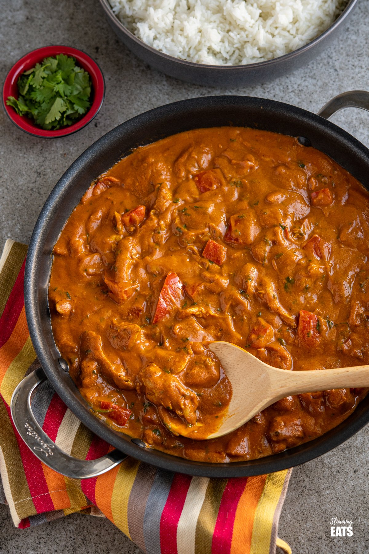 chicken curry in a black double handled frying pan with a wooden spoon, dark grey bowl of rice in the background