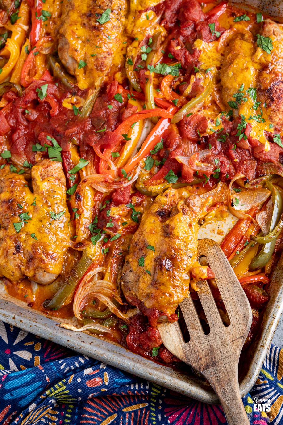 cheesy topped sausages with peppers, onion and salsa on baking tray with wooden spatula