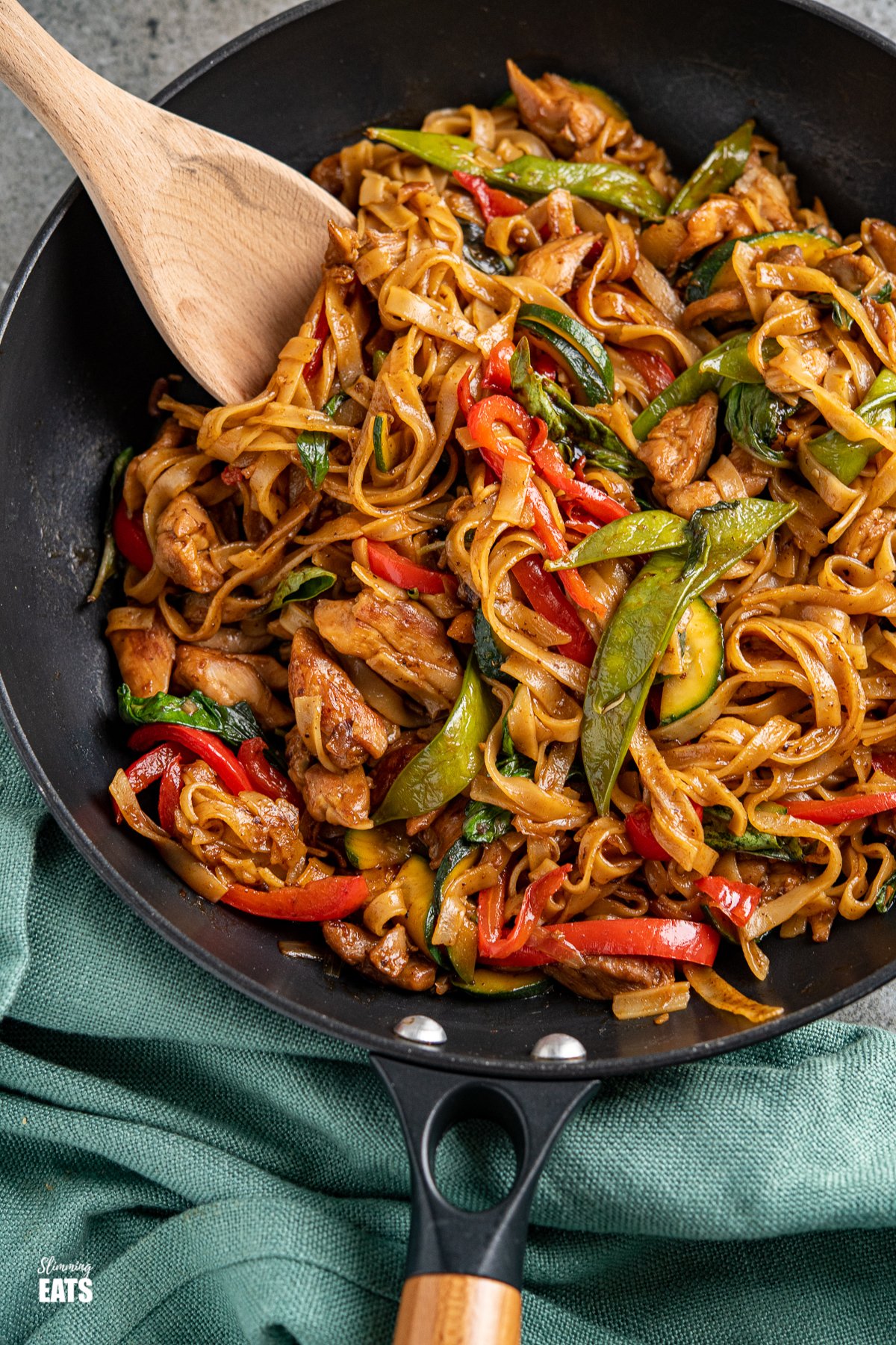 Drunken Chicken Noodles - love spicy noodle dishes? then this is the perfect recipe for you. Tender pieces of chicken with vegetables and rice noodles all coated in a flavoursome spicy sauce finished off with fragrant Thai Basil. drunken noodles - chicken, vegetables and rice noodles in a spicy flavoursome sauce in a black pan with a wooden handle