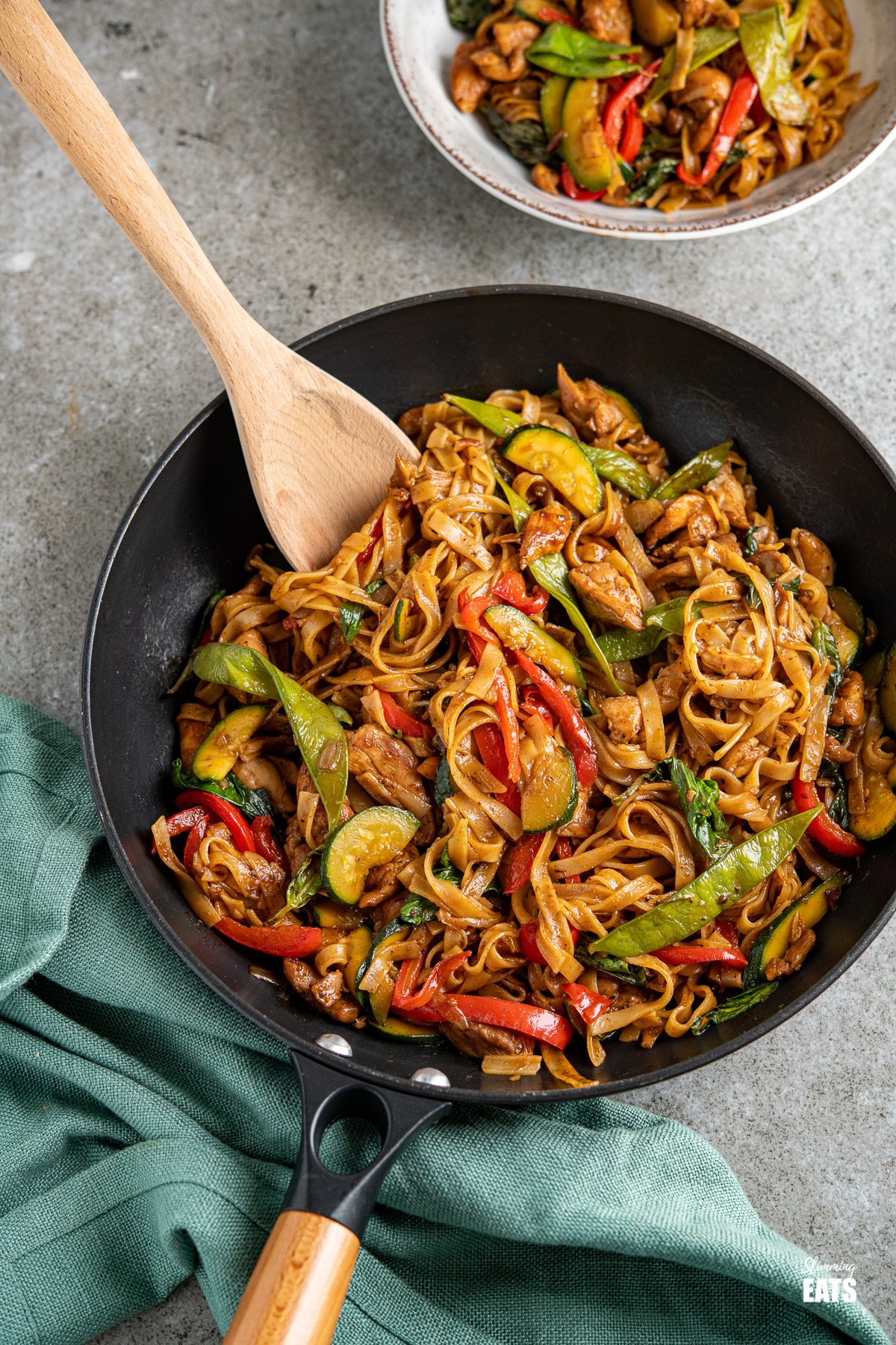 Drunken Chicken Noodles - love spicy noodle dishes? then this is the perfect recipe for you. Tender pieces of chicken with vegetables and rice noodles all coated in a flavoursome spicy sauce finished off with fragrant Thai Basil. drunken noodles - chicken, vegetables and rice noodles in a spicy flavoursome sauce in a black pan with a wooden handle