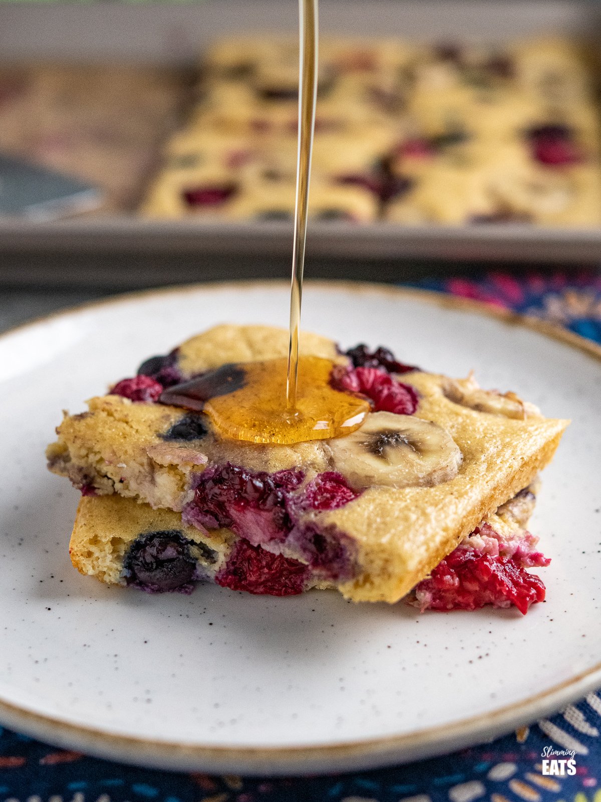 Sheet Pan Oat Pancakes - love pancakes but hate the flipping and standing at the stove? These easy sheet pan oat pancakes with berries and banana is the perfect effortless breakfast. Slimming World and Weight Watchers friendly Sheet Pan Oat Pancakes on a white plate with cream rim being drizzing with maple syrup, sheet pan in background