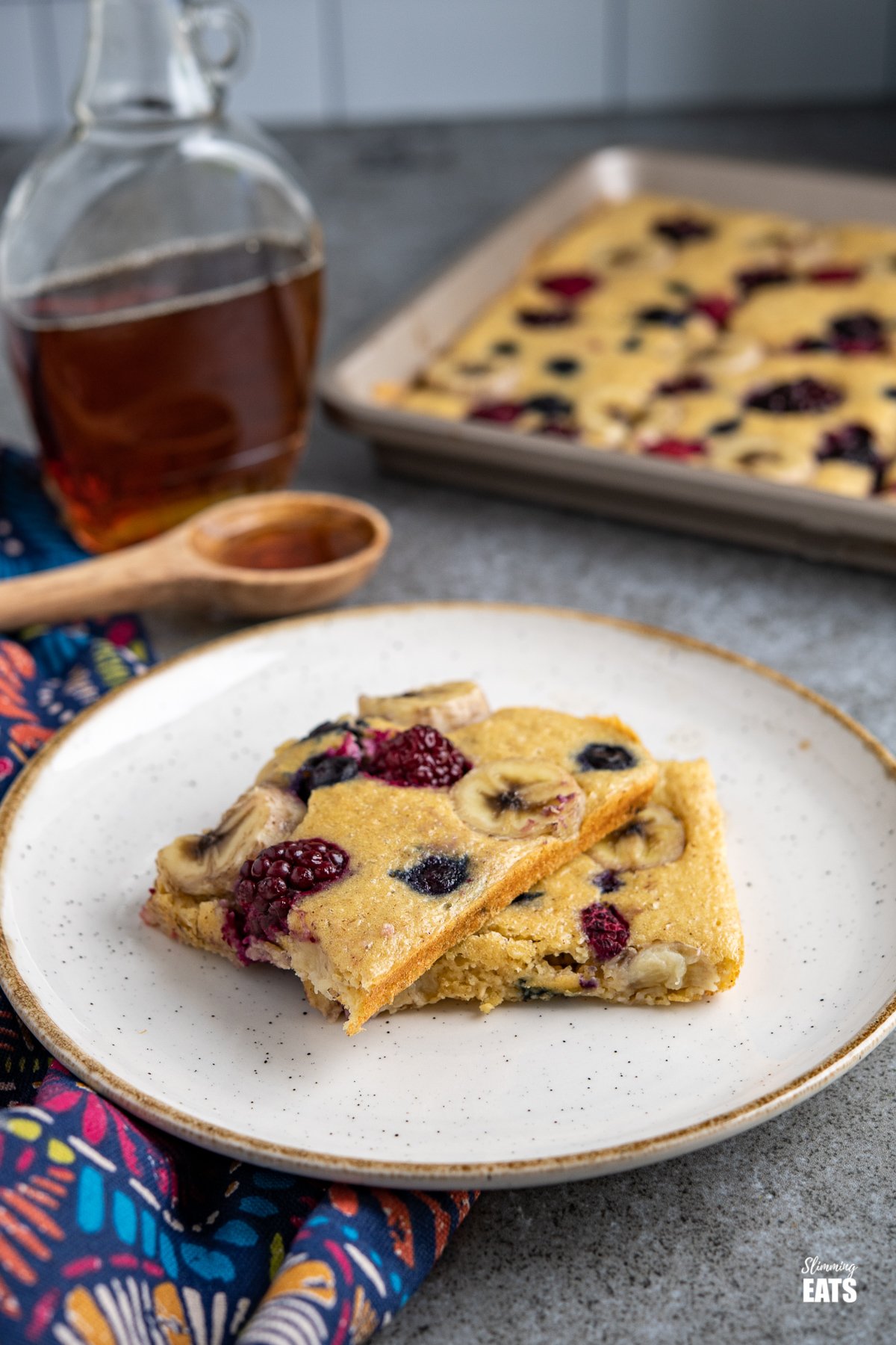 Sheet Pan Oat Pancakes - love pancakes but hate the flipping and standing at the stove? These easy sheet pan oat pancakes with berries and banana is the perfect effortless breakfast. Slimming World and Weight Watchers friendly Sheet Pan Oat Pancakes on a white plate with cream rim, maple syrup and sheet pan in background