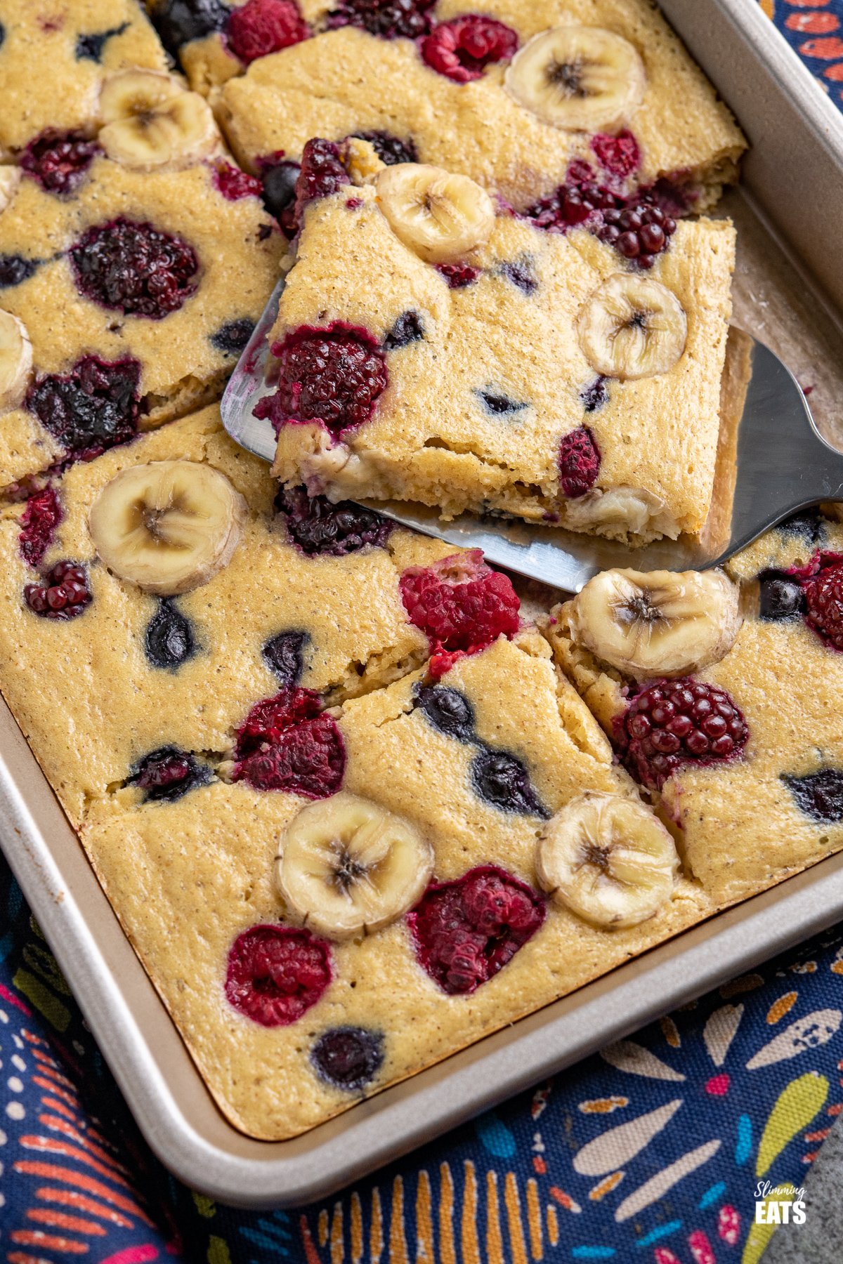 Sheet Pan Oat Pancakes - love pancakes but hate the flipping and standing at the stove? These easy sheet pan oat pancakes with berries and banana is the perfect effortless breakfast. Slimming World and Weight Watchers friendly sliced Sheet Pan Oat Pancakes in a baking tray with a slice on a spatula