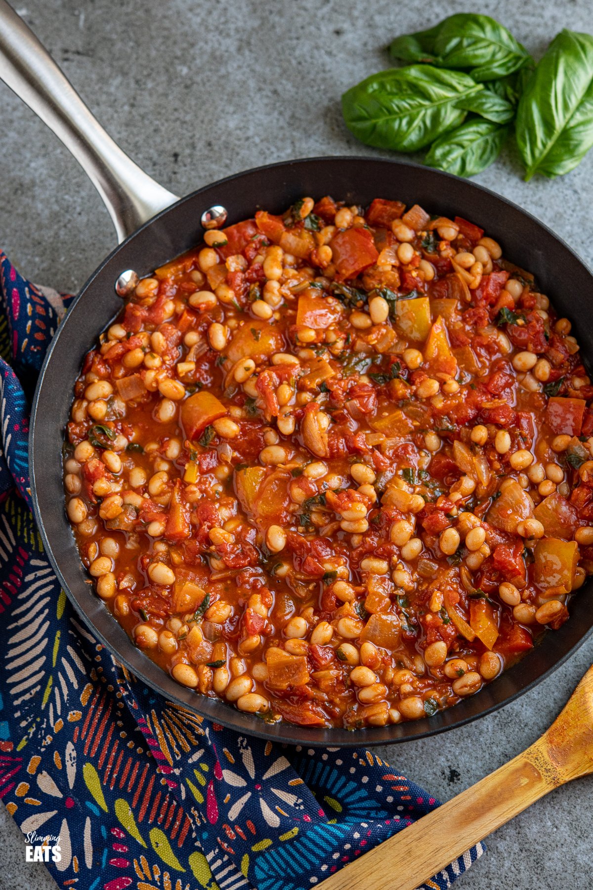 Tomatoes and White Beans in a black frying pan with a bunch of fresh basil in top right corner. 