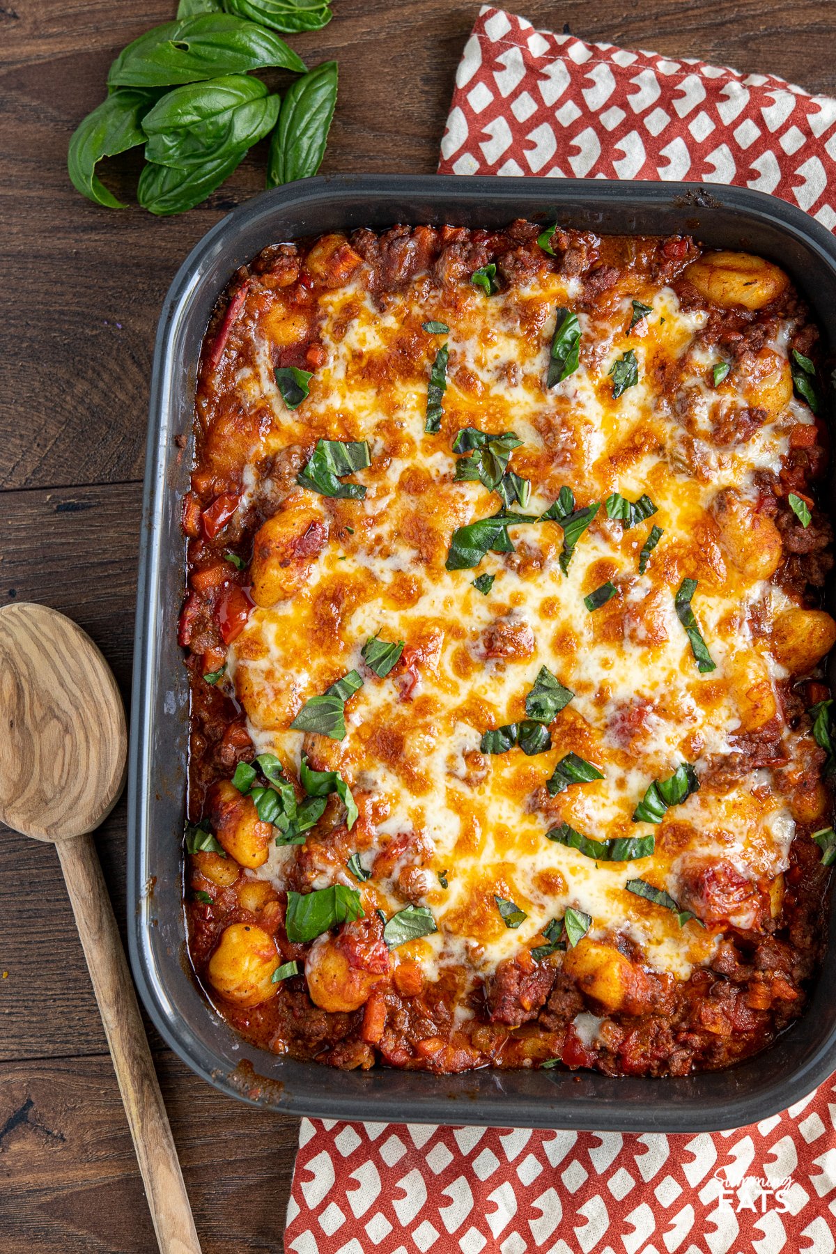 cheesy bolognese gnocchi bake in a grey baking dish with scattered basil leaves and olive wood spoon to the left