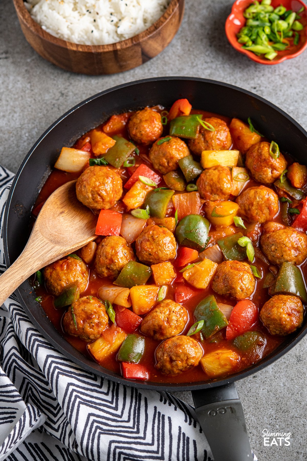 sweet and sour chicken meatballs in a black frying pan with vegetables, wooden bowl of jasmine rice in background and terracotta pinch bowl with spring onions