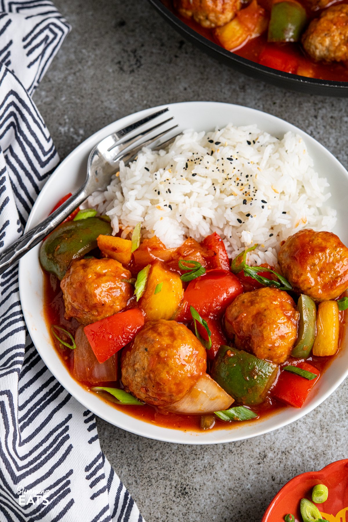 sweet and sour chicken meatballs in white bowl with jasmine rice, fork placed to the left with a black and white napkin to the side. 