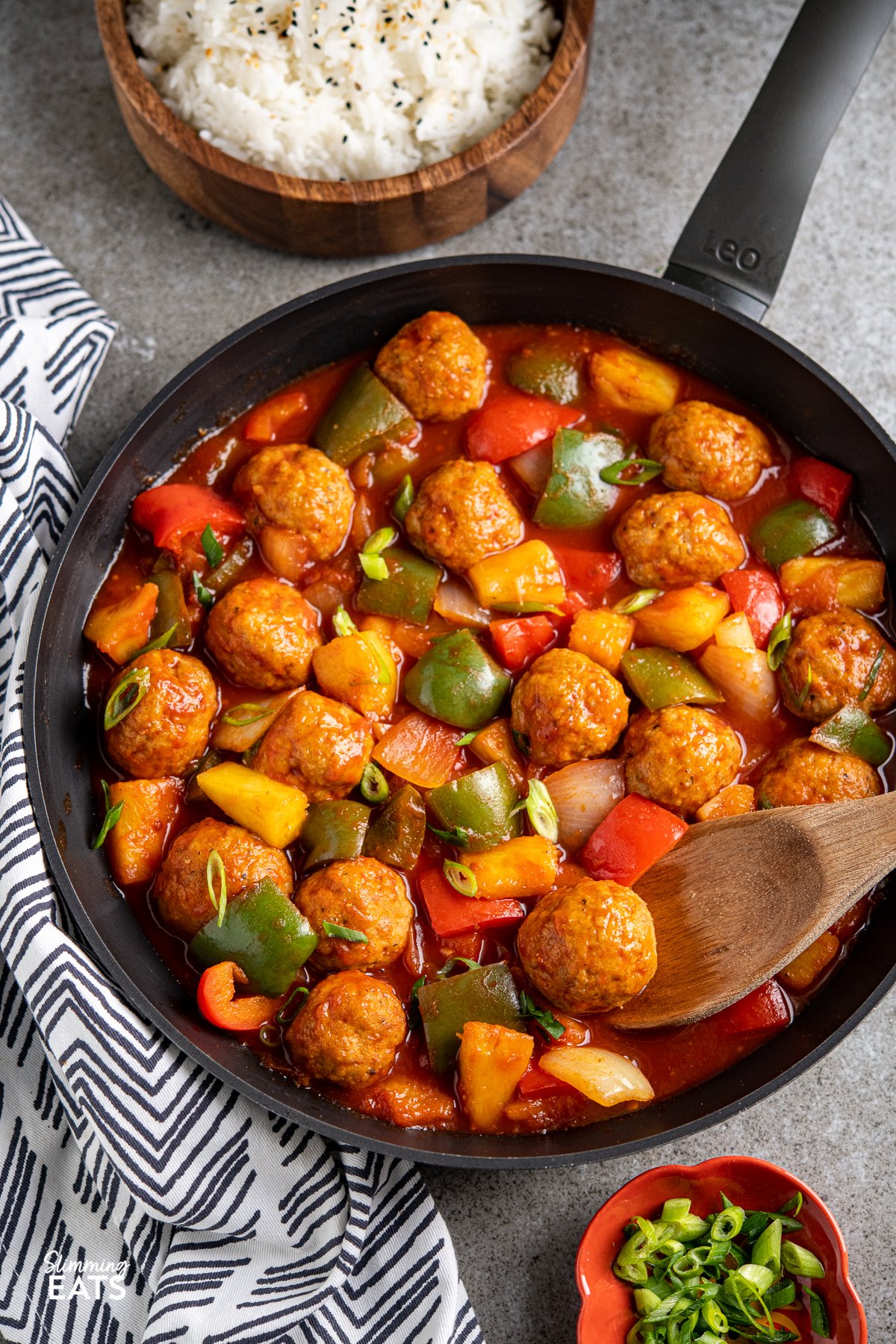 sweet and sour chicken meatballs in a black frying pan with vegetables and a wooden bowl of jasmine rice in background