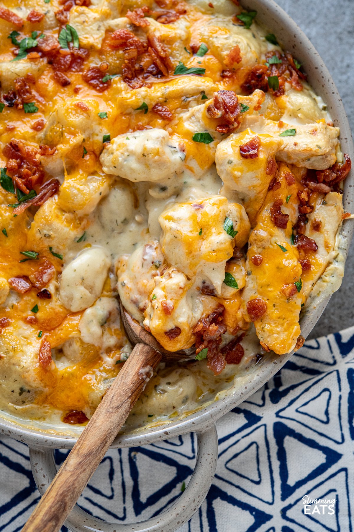 close up of crack chicken gnocchi  on olive wooden spoon in double handled grey brown ceramic dish with melted cheddar and crisp golden bacon.