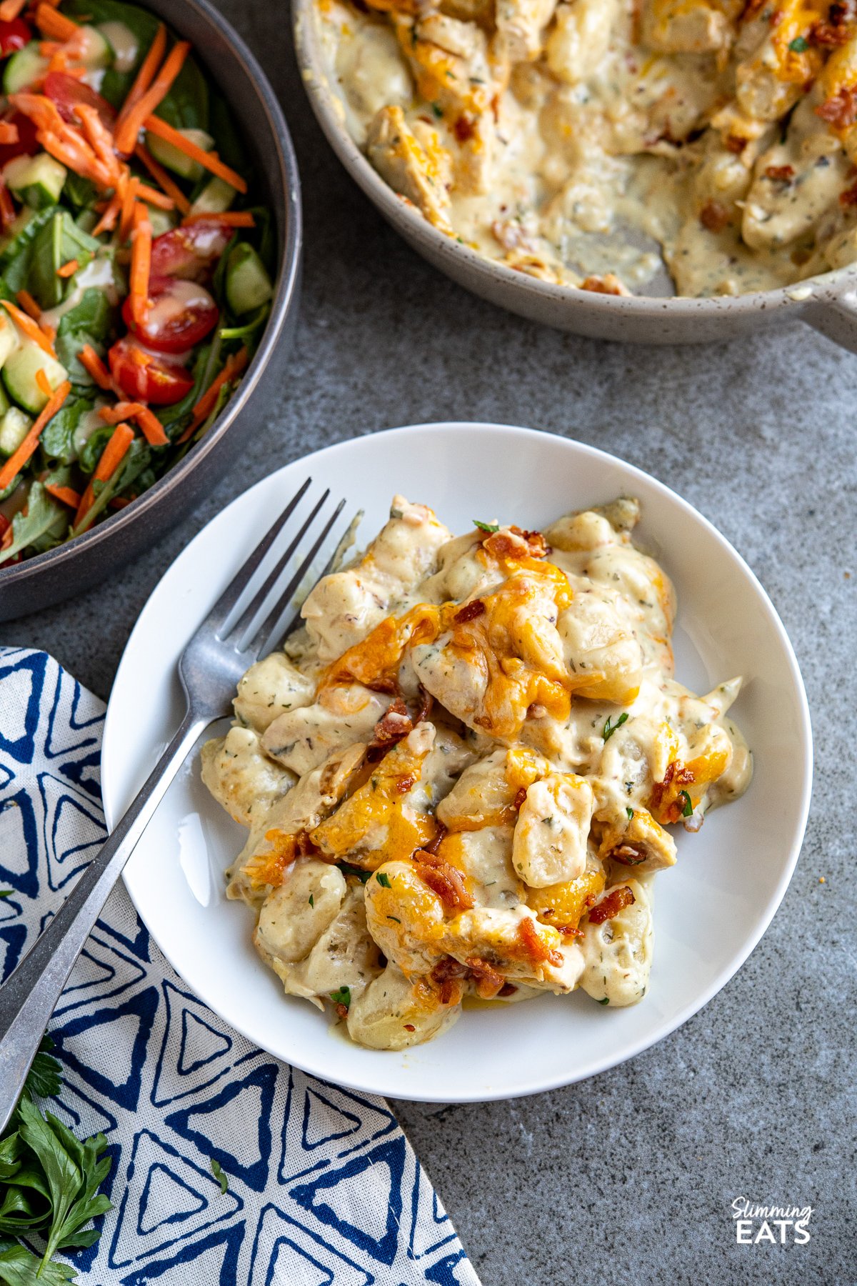 Crack Chicken Gnocchi - delicious tender pillows of gnocchi in a creamy delicious ranch style sauce with juicy chicken breast, melted cheddar and golden bacon. The only regret you will have is not making this dish. Slimming Eats and Weight Watchers friendly Crack Chicken Gnocchi in white bowl with fork, dish of gnocchi in background and grey bowl with salad.