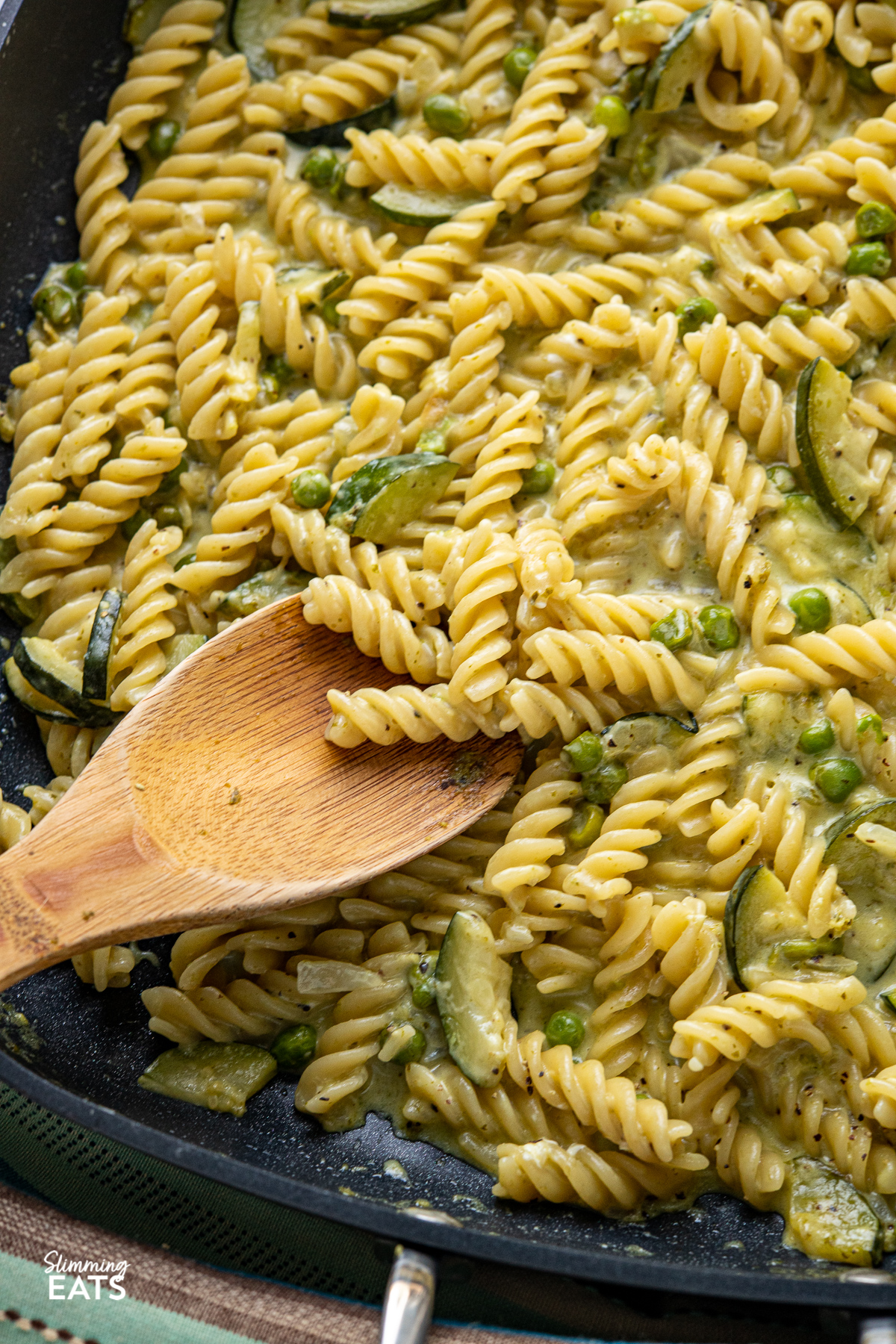 close up of One Pot Creamy Pesto Courgette Pea Pasta in black all-clad skillet with wooden spoon