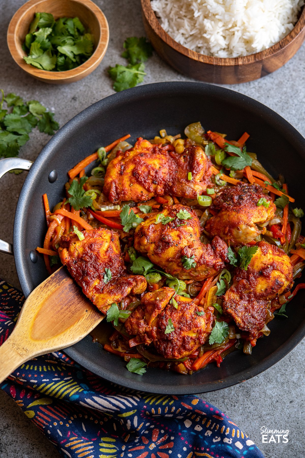 cooked fusion marinated chicken thighs over a bed of vegetables in a two handled frying pan with wooden bowl of rice and cilantro in background