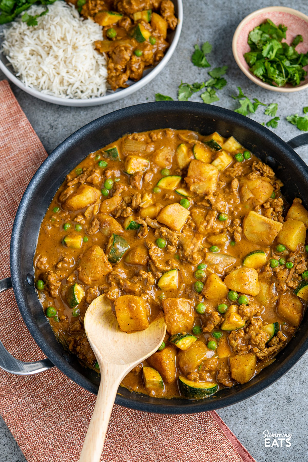 ground turkey and potato curry in black double handled frying pan with serving on a plate in backround wiht rice and small bowl of fresh coriander