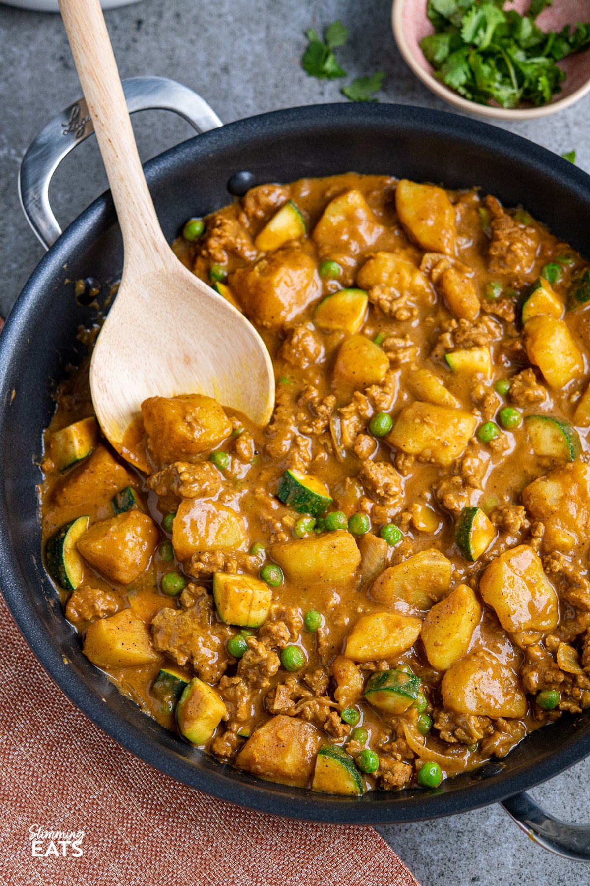 ground turkey and potato curry with wooden curry spoon in a black double handled frying pan, small bowl of chopped coriander in background. 