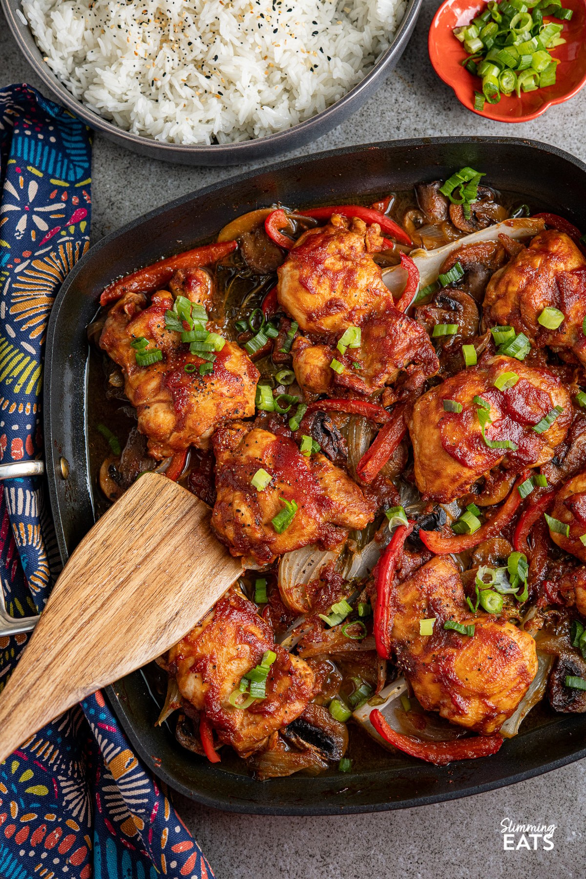 Sriracha Peach Chicken Bake with vegetables in square All-Clad skillet with grey bowl of rice and orange pinch bowl with sliced spring onions