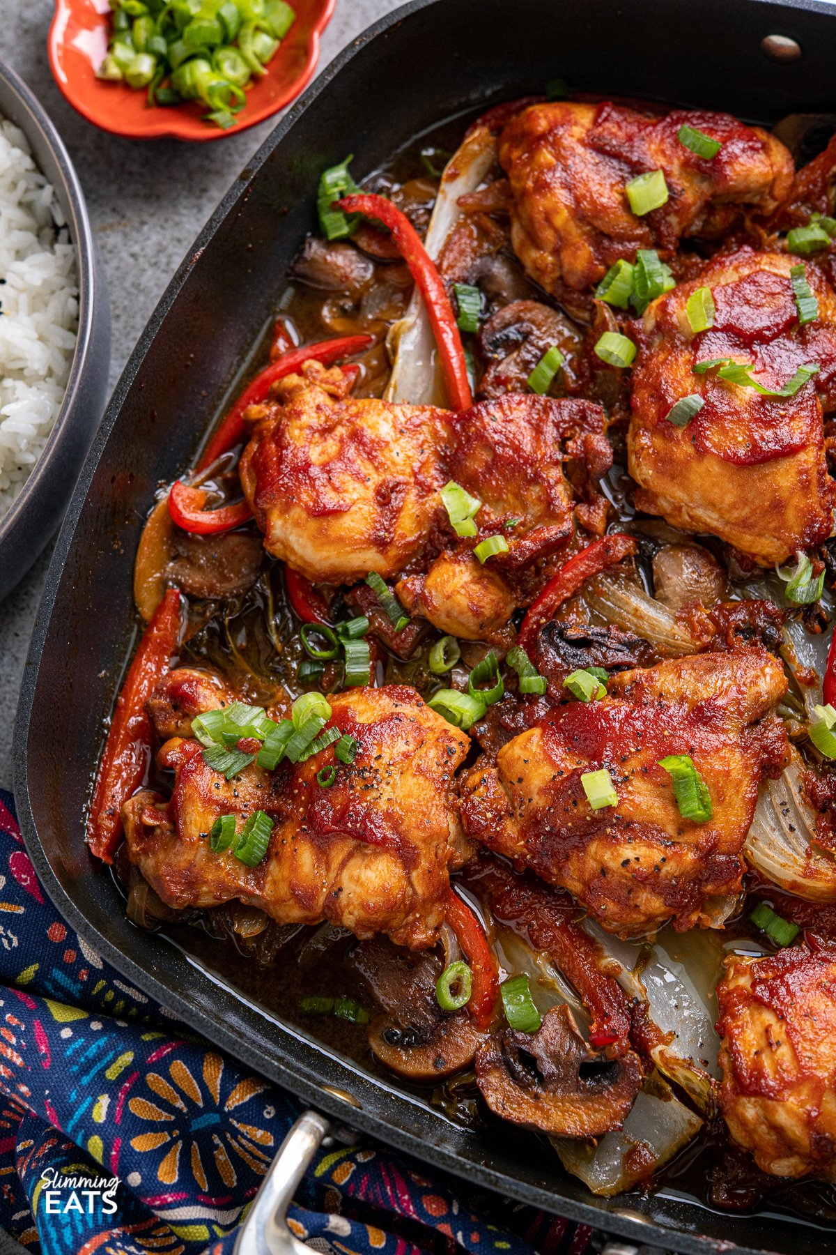 close up of Sriracha Peach Chicken Bake with vegetables in square All-Clad skillet  with dark grey bowl of rice and pinch bowl with sliced spring onions