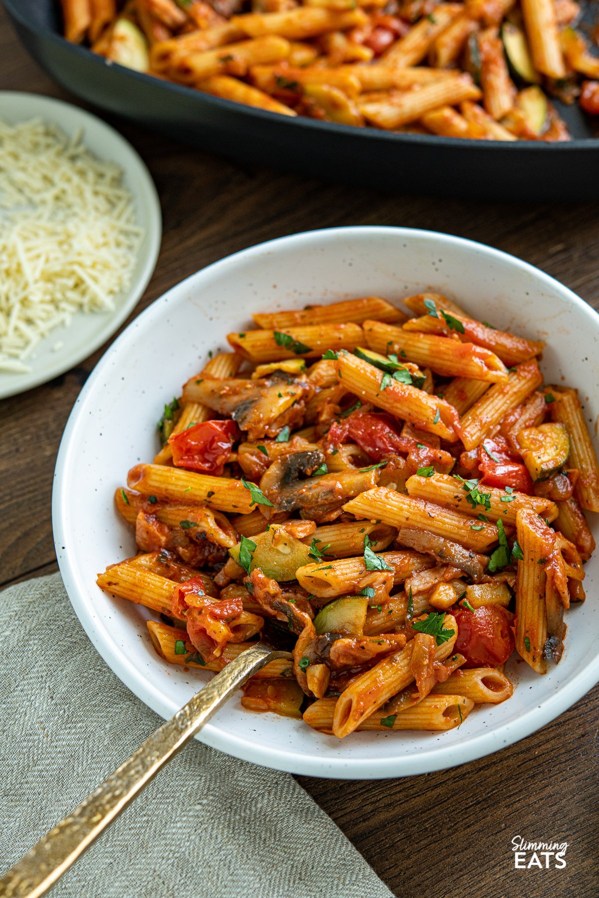 Mushroom, Bacon, Tomato and Zucchini Pasta in white bowl, with plate of parmesan and all-clad square skillet in background