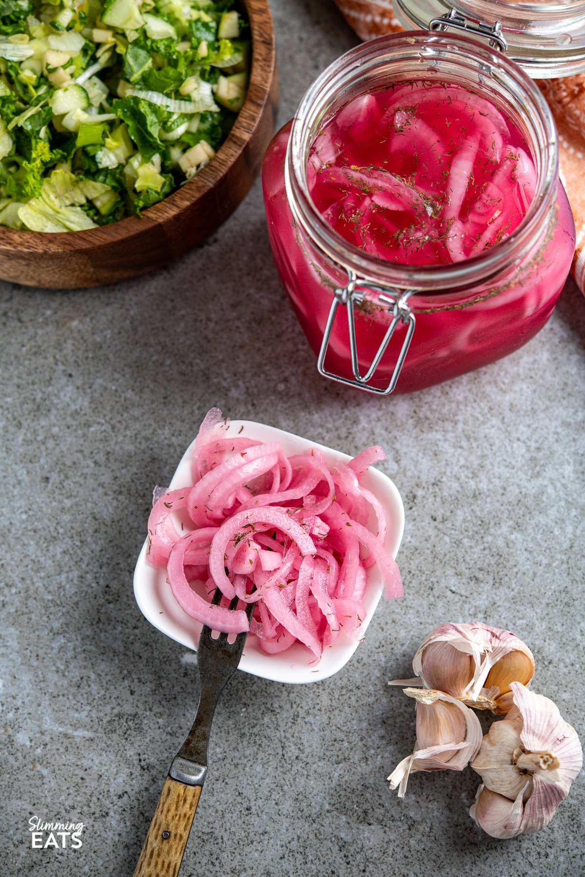 Homemade Pickled Red Onions in a small white square bowl with fork, jar of pickled onions in background and wooden bowl with salad, bulb of garlic at below bowl