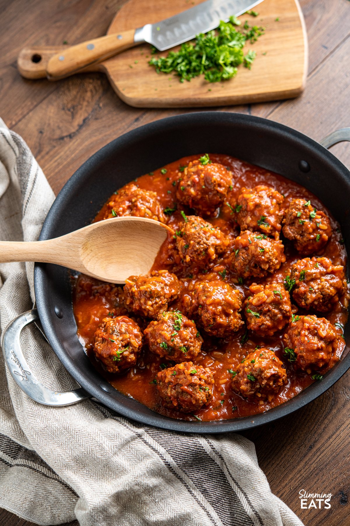 Porcupine Meatballs - Tender beef meatballs with rice in a rich and delicious tomato sauce - A perfect recipe for the whole family. Slimming Eats and Weight Watchers friendly Porcupine Meatballs with wooden spoon in black skillet, chopping board with parsley in background.