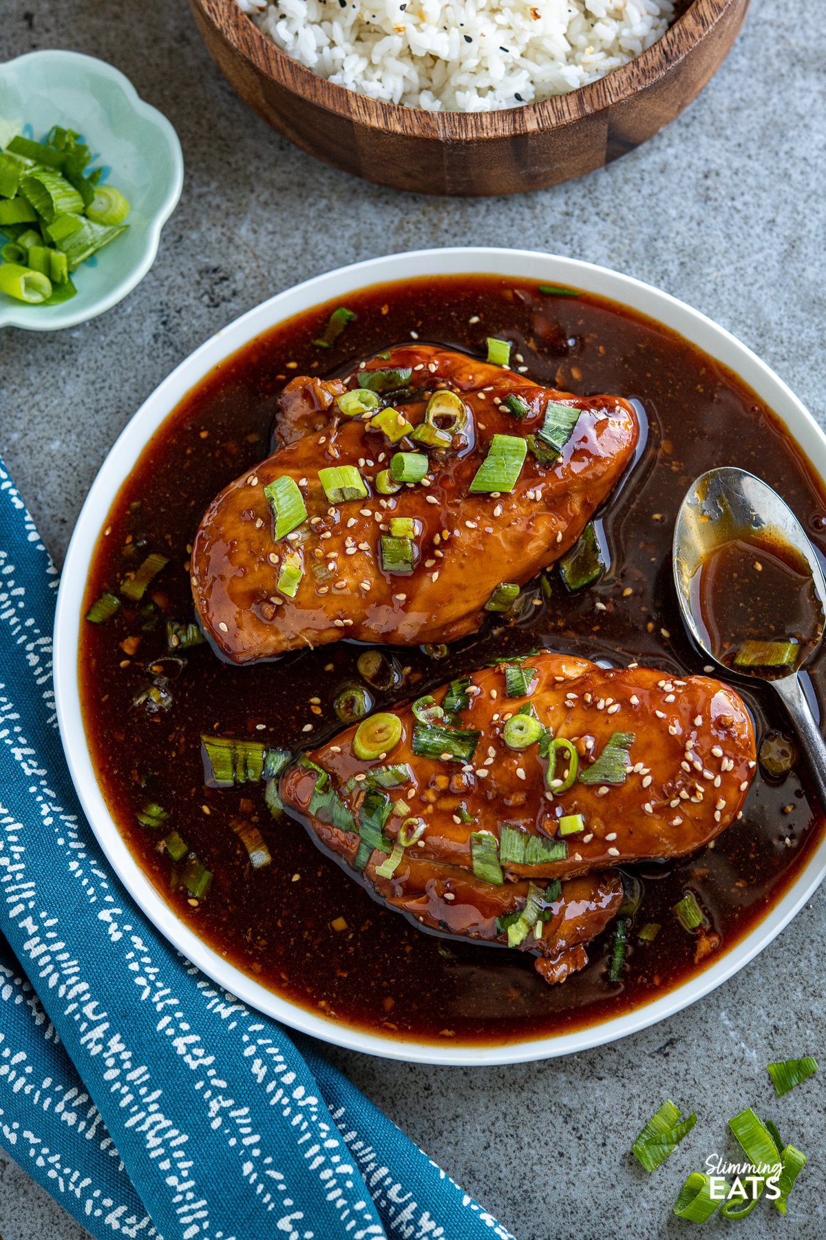 Slow Cooker Teriyaki Chicken on white plate with scattered spring onions and sesame seeds and a blue and white towel to left. 