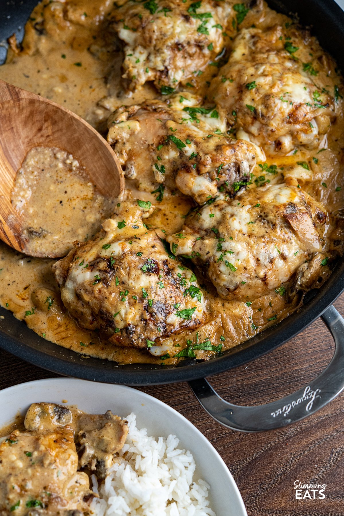 smothered creamy garlic chicken with mushrooms in black double handled frying pan with white bowl with serving off to side.