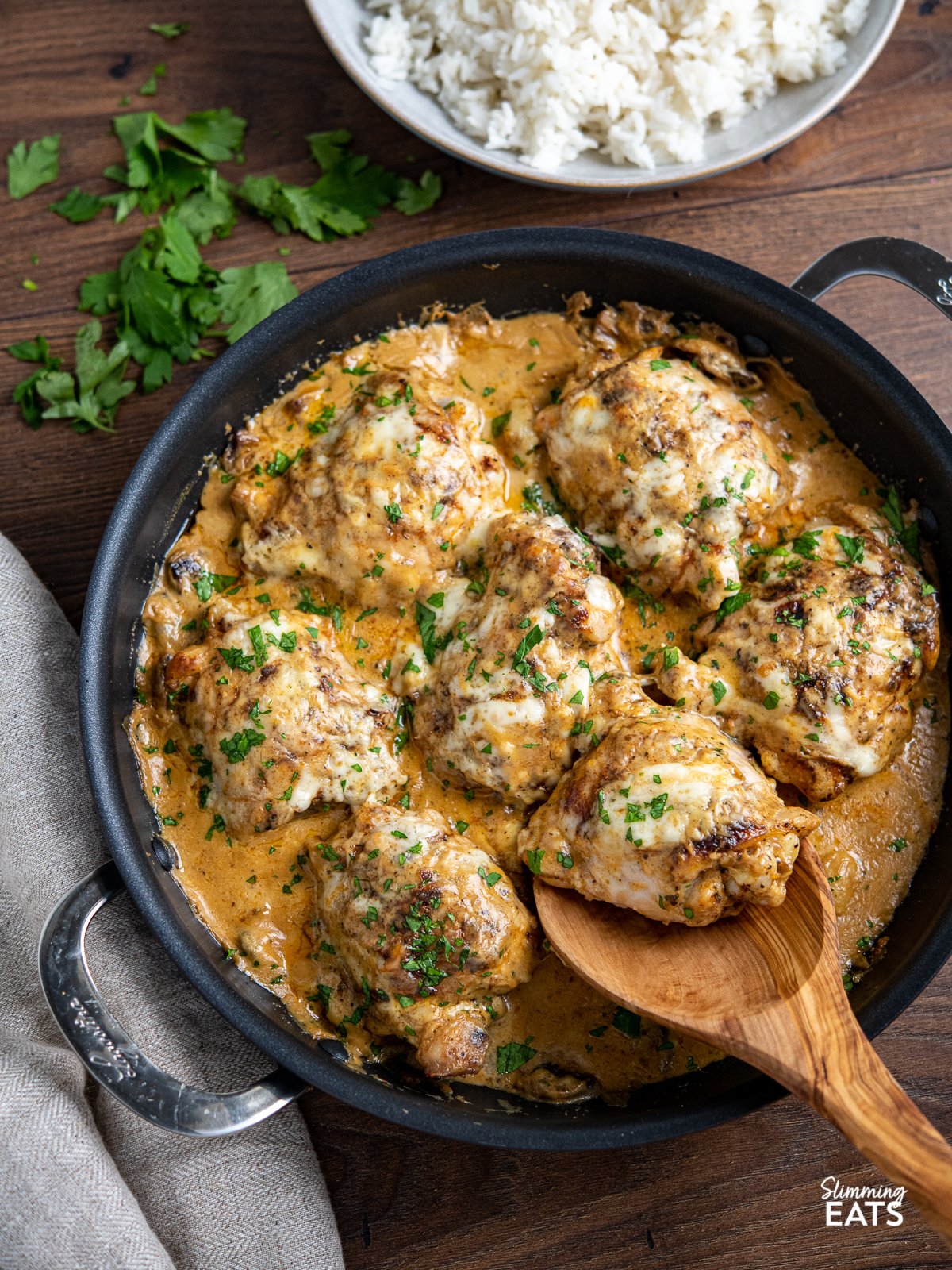 smothered creamy garlic chicken with mushrooms in black double handled frying pan  with olive wood spoon scooping up one chicken thigh. 