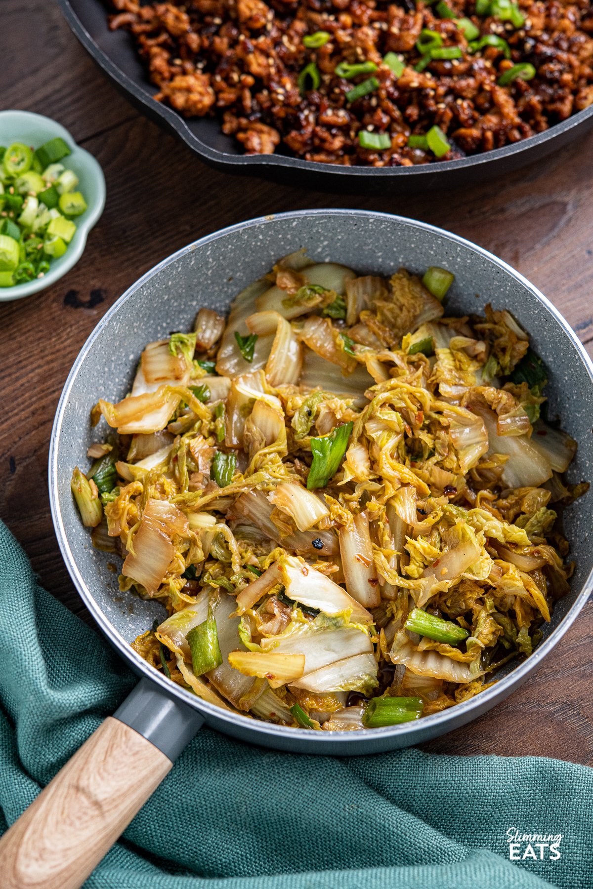 Spicy Garlic Napa Cabbage in grey frying pan with wooden handle, pan of Korean ground pork in background