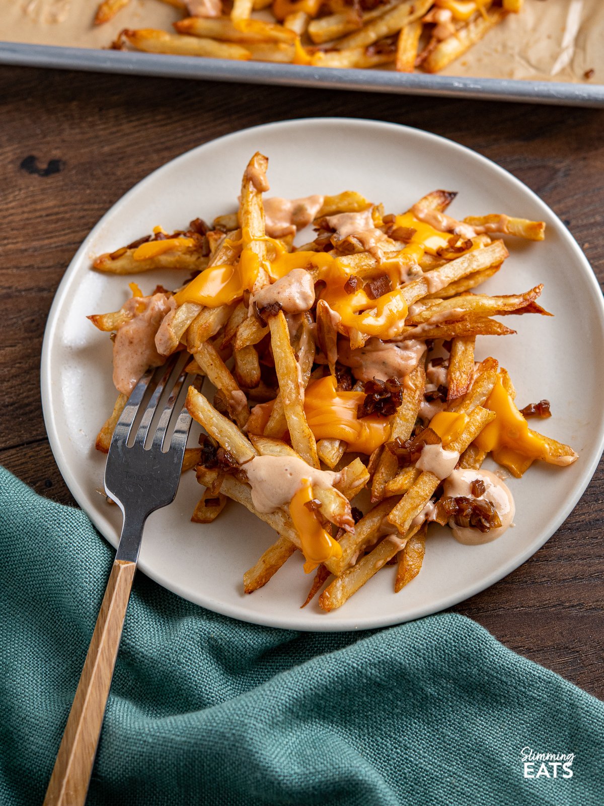 Indulge with these classic fakeaway Animal Style Fries - sweet sauteed onions, golden oven baked fries with melted American cheese and a classic special sauce what more could one want? plate of Copycat Oven Baked Animal Style Fries on a cream plate, for place on side, baking tray in background.