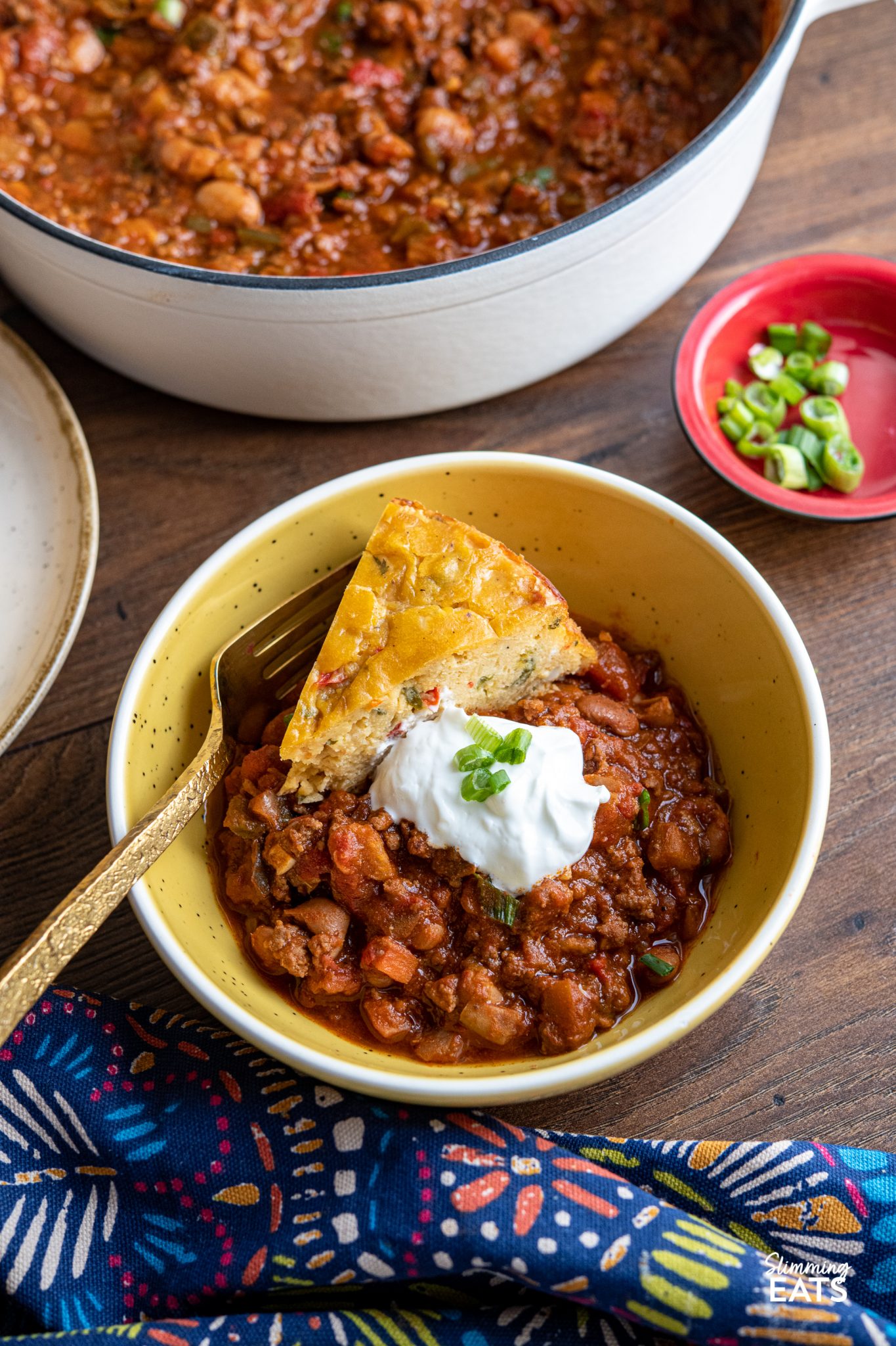 beef and pinto bean chilli in a small yellow bowl with slice of  lentil cheddar bake and sour cream with spring onions