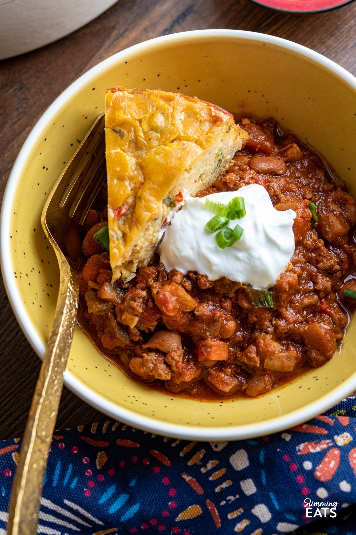 close up of beef and pinto bean chilli in a small yellow bowl with slice of  lentil cheddar bake and sour cream with spring onions