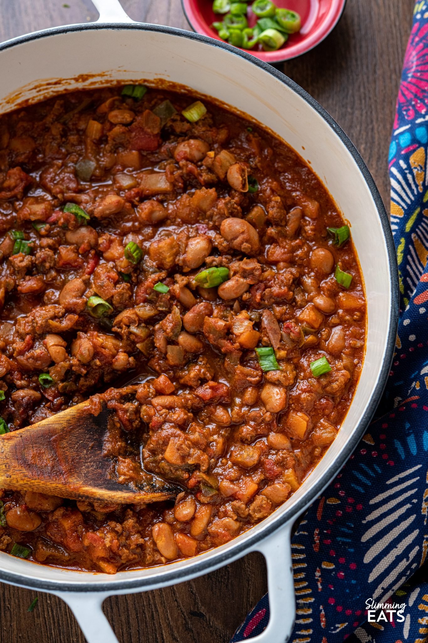 close up of beef and pinto bean chilli in a white cast iron pan with small red bowl of chopped spring onion