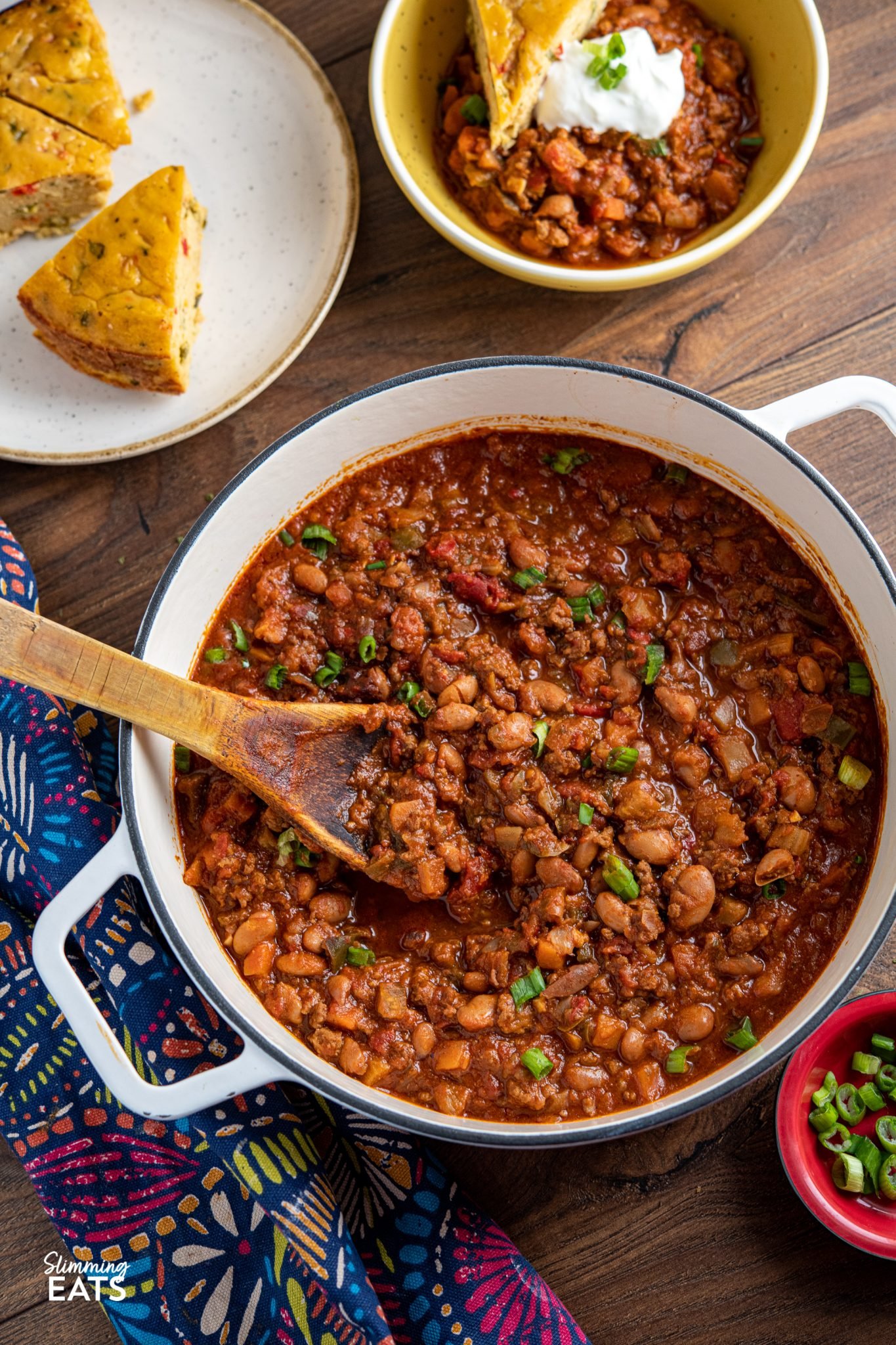 beef and pinto bean chilli in a white cast iron pan with bowl of chilli in background with plate with lentil cheddar bake