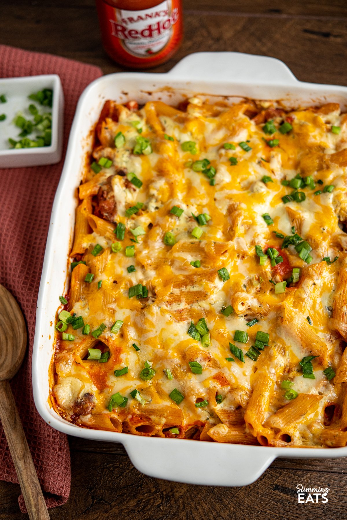 Buffalo Chicken Pasta Bake in white baking dish with chopped spring onions in a square dish and Frank's Red Hot Sauce in background