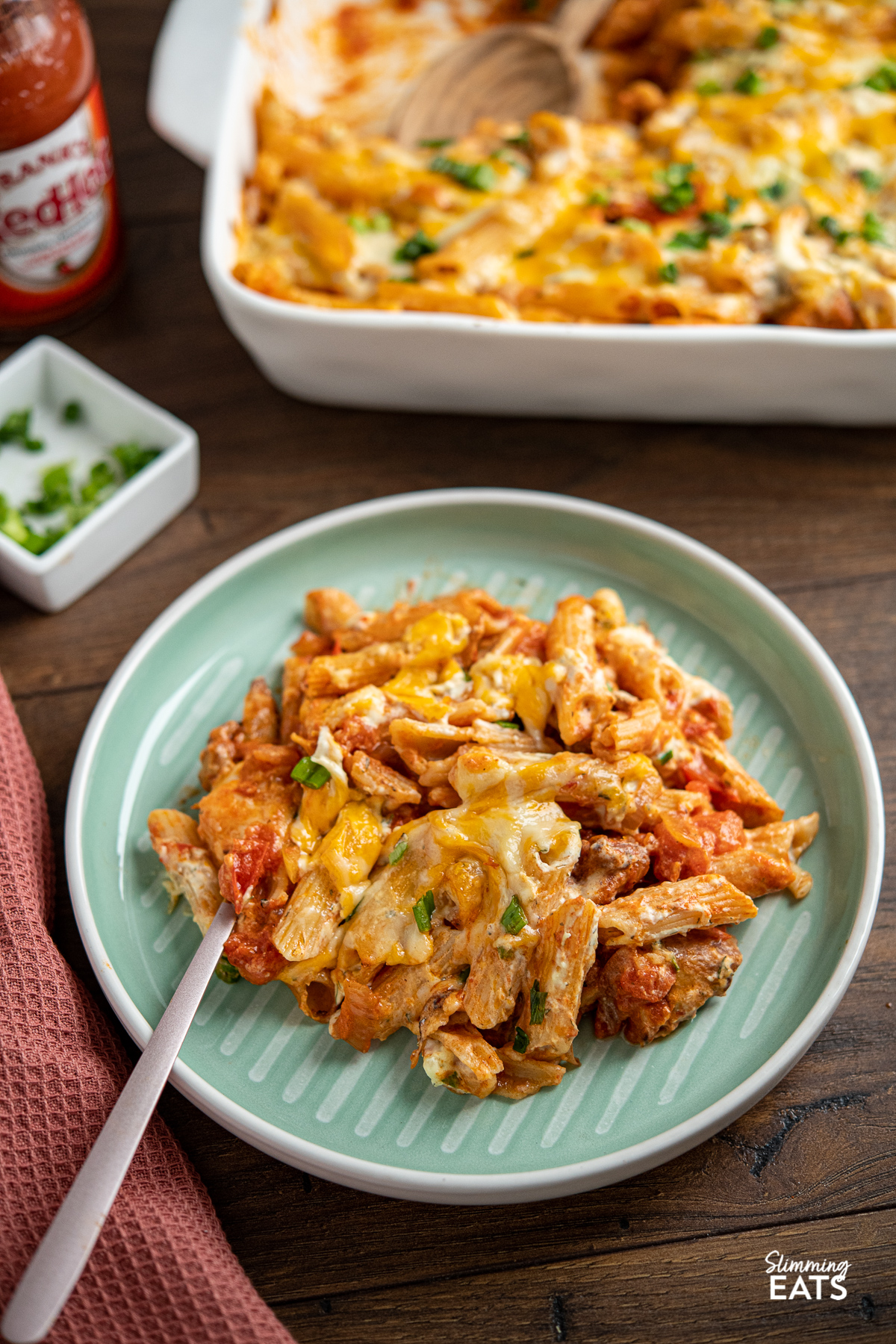 A serving of Buffalo Chicken Pasta Bake on a turquoise plate with fork, white baking dish with pasta bake in background and Frank's Red Hot Sauce