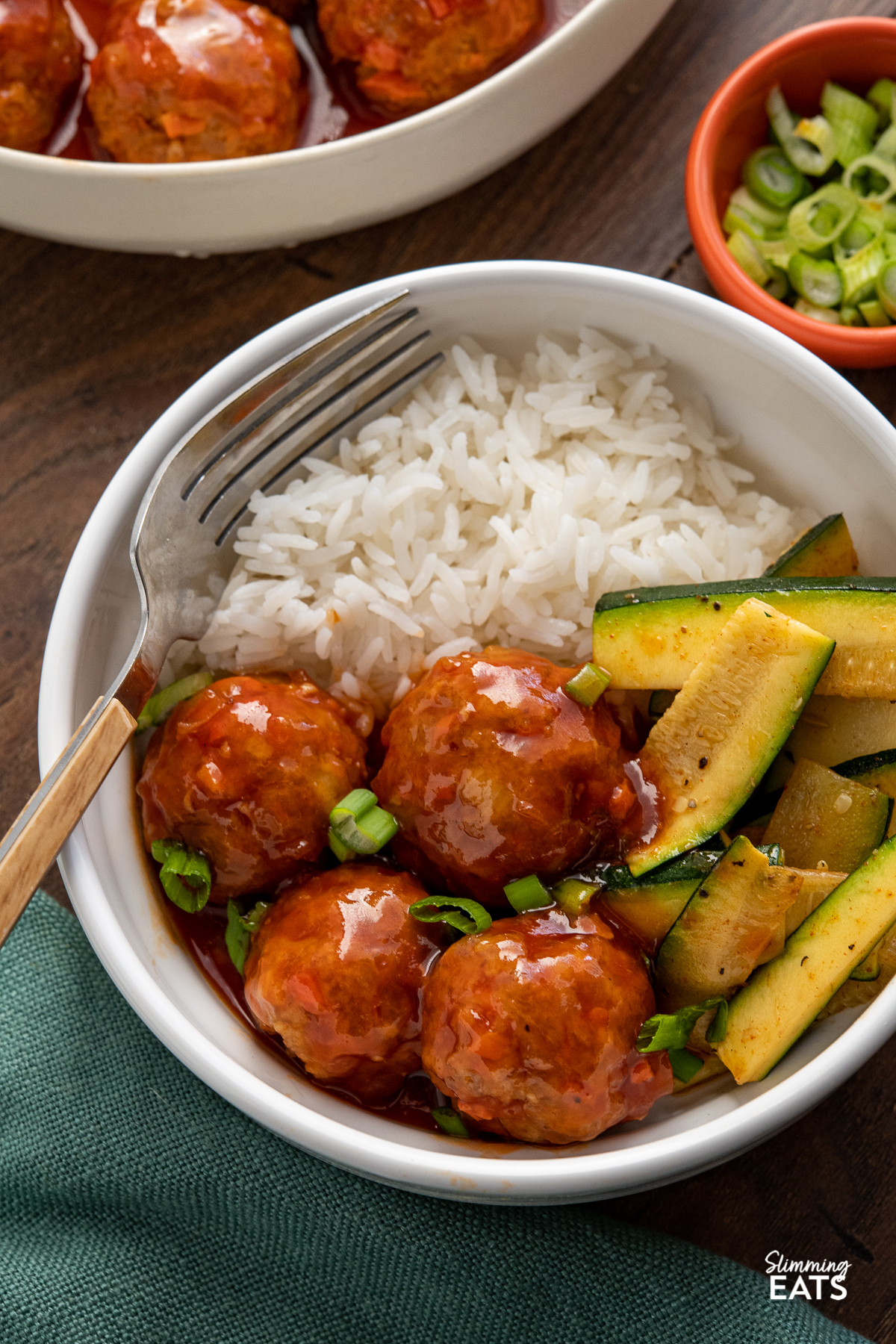 buffalo chicken meatballs in white bowl with rice and sauteed zucchini, bowl of meatballs and small orange bowl of chopped green onions in background