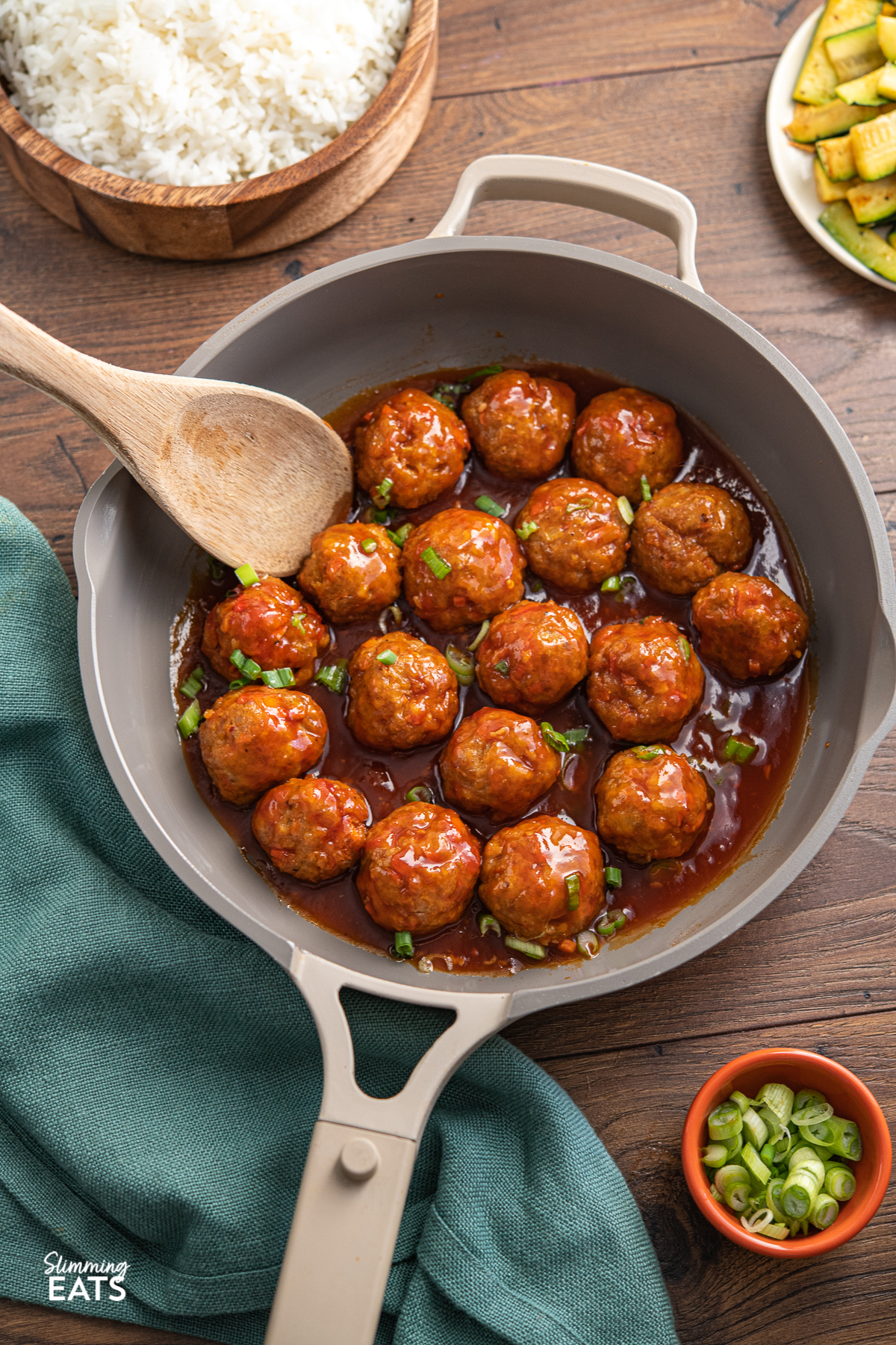 buffalo chicken meatballs in a beige  non stick always pan with wooden spoon place in pan and wooden bowl of rice in background, turquoise fabric placed to left