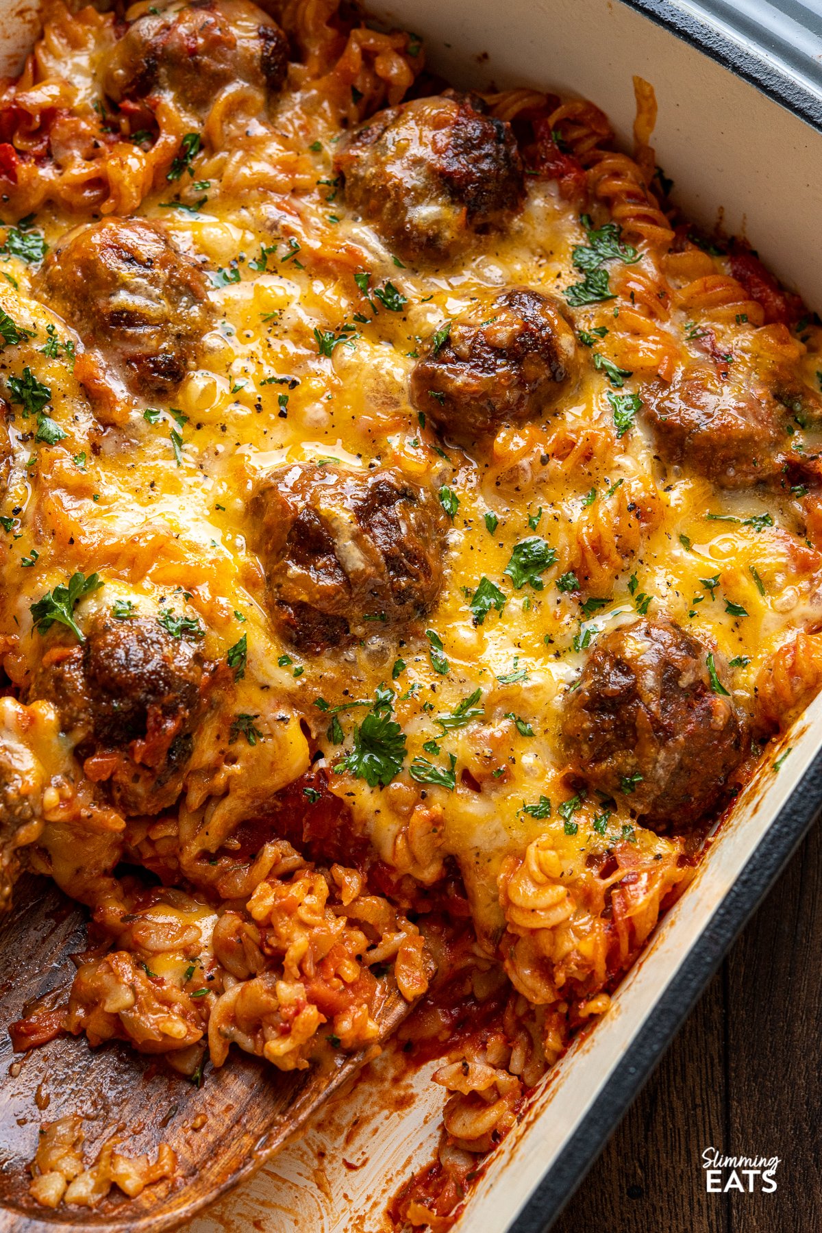 Cheesy Meatball Pasta Bake - delicious and lean homemade beef meatballs in a rich tomato sauce all stirred into fusilli pasta, topped with cheese and baked in the oven until melted and golden for a perfect family meal. wooden spatula spoon up meatball pasta bake. from a baking dish