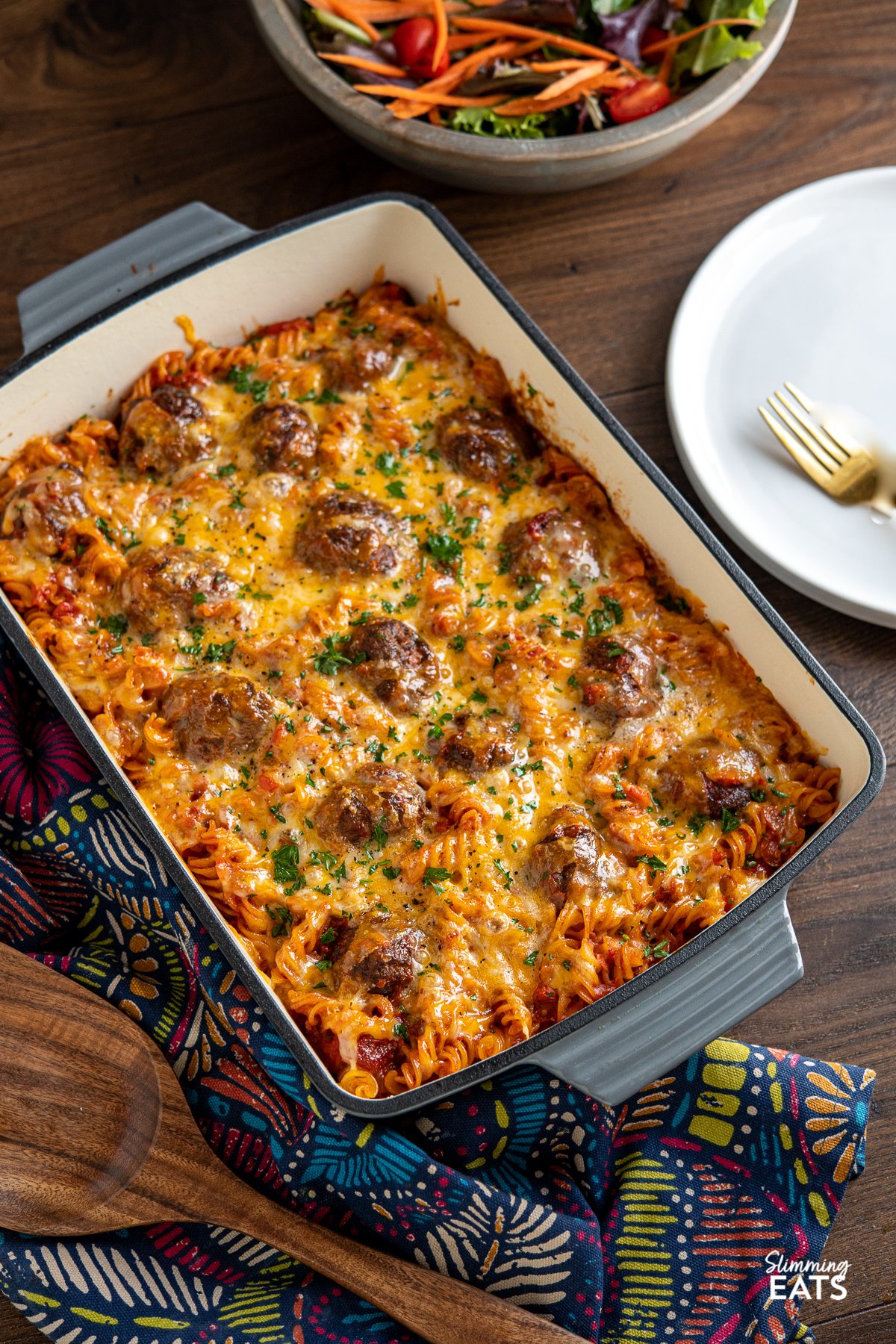 Cheesy Meatball Pasta Bake in baking dish with white plate to the left and wooden spatula below. 