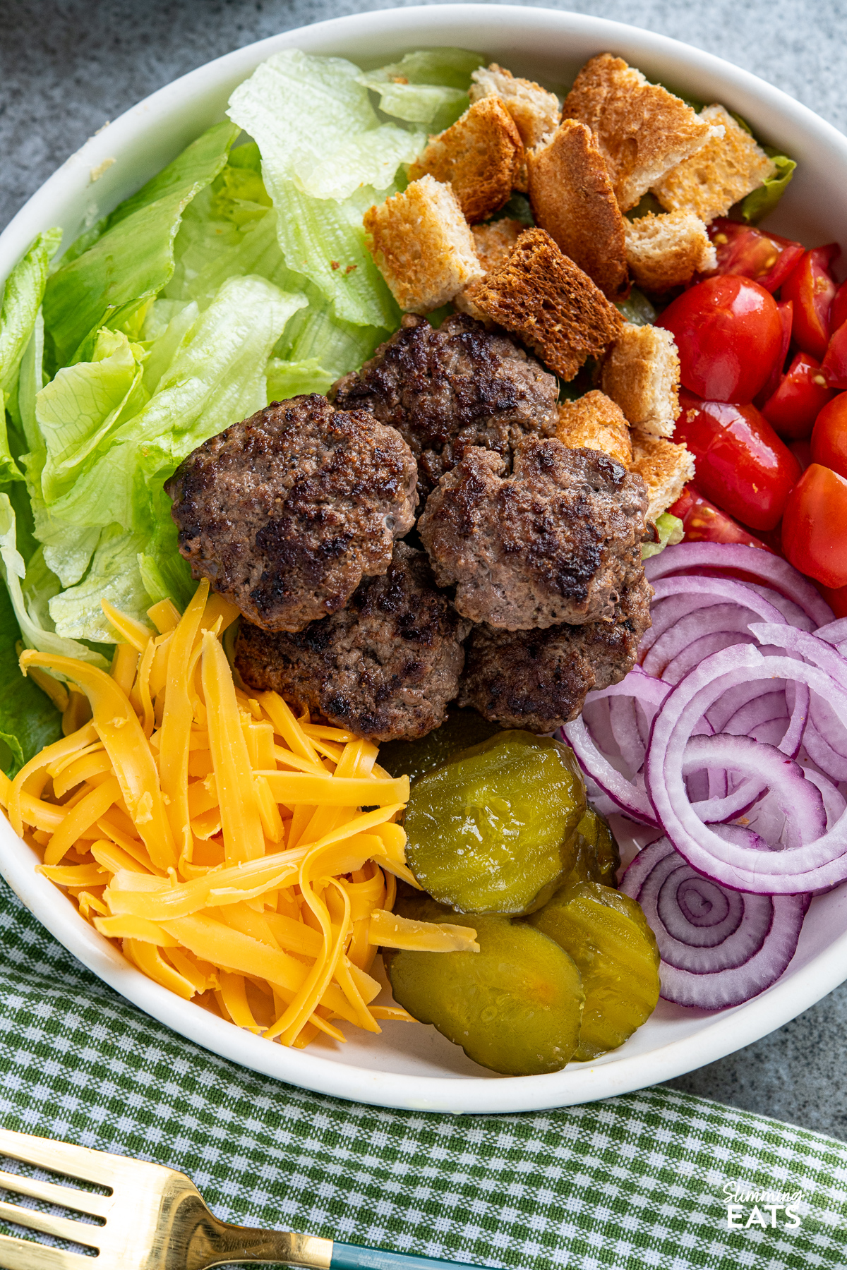 close up of cheeseburger salad in a white bowl with dressing and croutons placed next to bowl