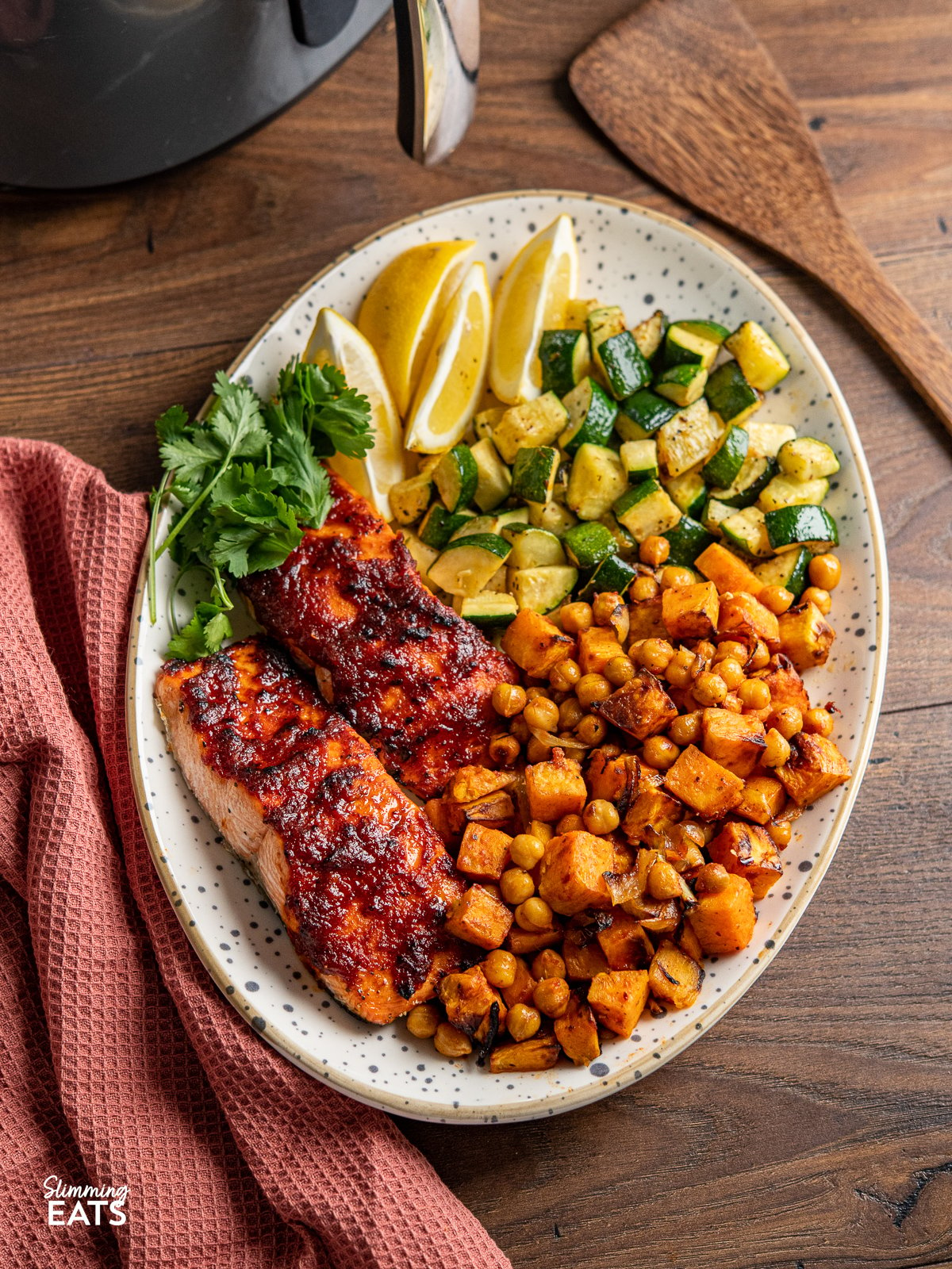Air Fryer Honey Harissa Salmon on a oval spotted plate with sweet potato, chickpeas, zucchini, lemon wedges and coriander, wooden spoon and air fryer in background. 