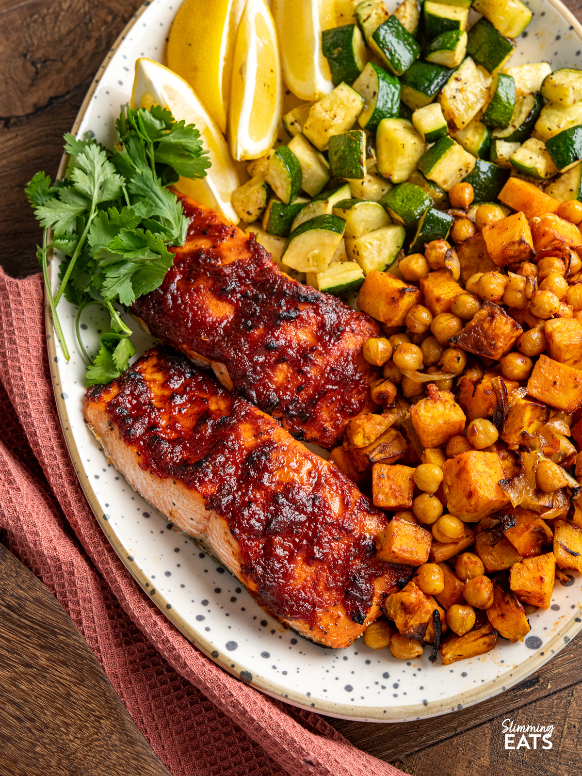 close up of Air Fryer Honey Harissa Salmon on a oval spotted plate with sweet potato, chickpeas, zucchini, lemon wedges and coriander,
