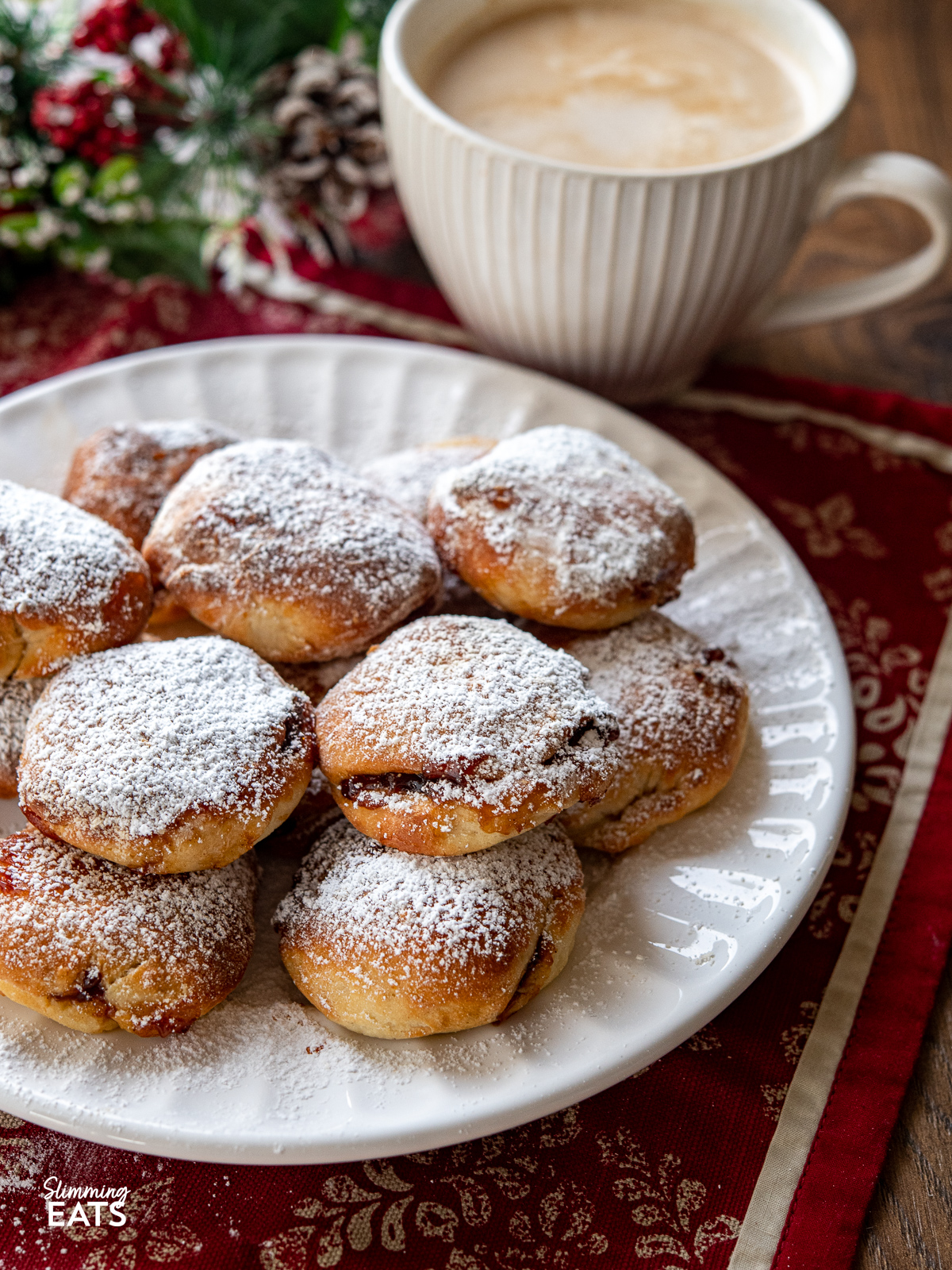 Air Fryer Mince Pie Bites on a white plate dusted with icing sugar with mug of coffee and christmas decorations in background