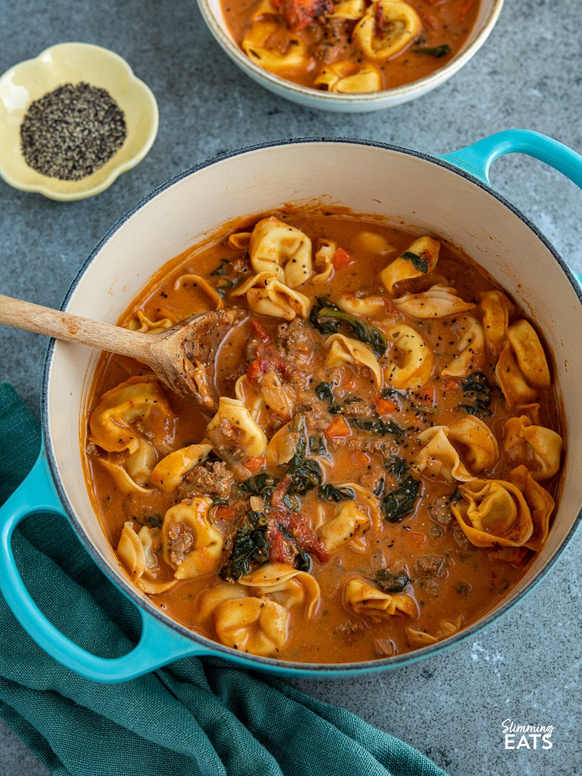 Creamy Italian Sausage Tortellini Soup in teal cast iron le creuset with bowl of soup and little flower bowl of black pepper in background