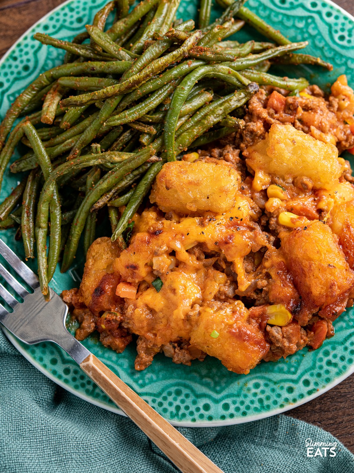 close up of Tater Tot Casserole served up on a turquoise plate with roasted green beans, fork placed diagonally at front of plate