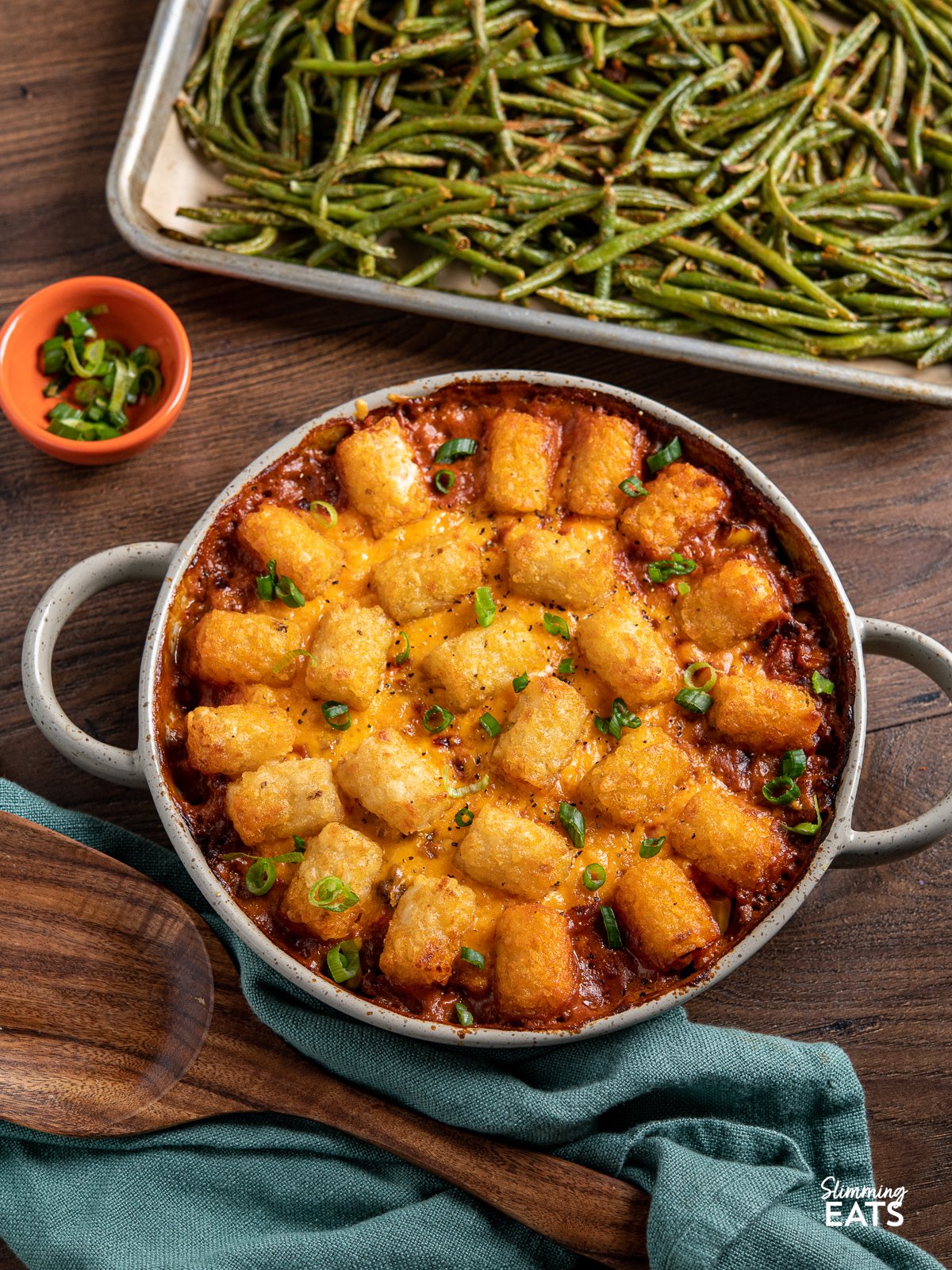Tater Tot Casserole is the perfect comforting family friendly dish - all made low calorie friendly with lean seasoned ground beef with carrots and corn in a creamy sauce, topped with melted cheese and golden crispy tater tots Tater Tot Casserole in a round oven dish with two handles, tray of roasted green beans in the background