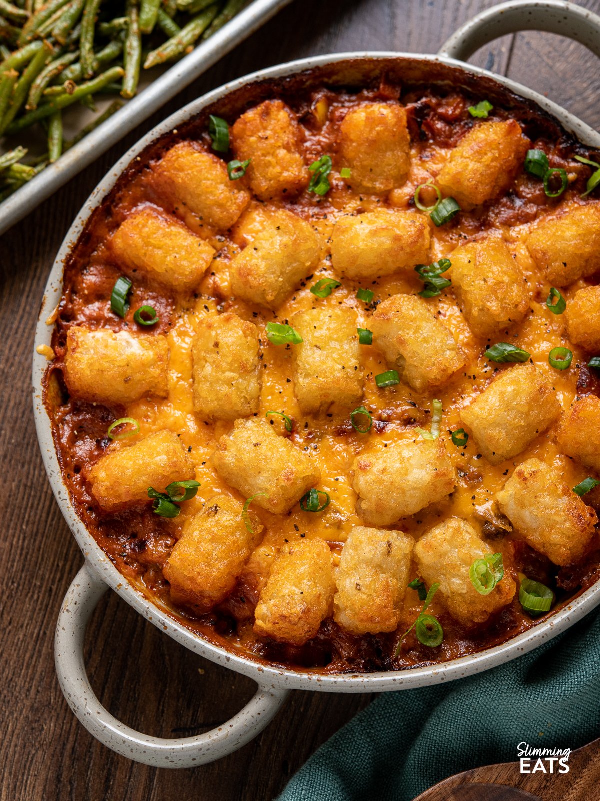 Tater Tot Casserole is the perfect comforting family friendly dish - all made low calorie friendly with lean seasoned ground beef with carrots and corn in a creamy sauce, topped with melted cheese and golden crispy tater tots close up of Tater Tot Casserole in a round oven dish with two handles, tray of roasted green beans in the background