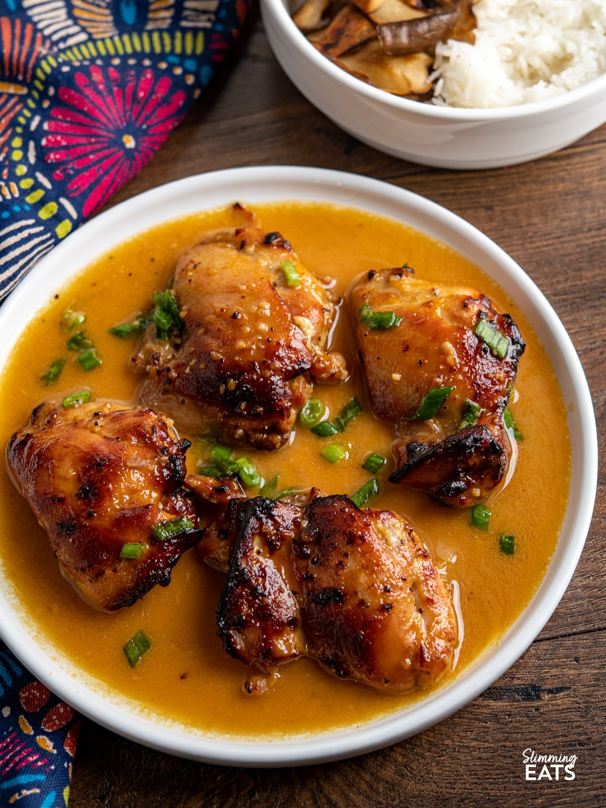 A simple air fryer recipe of tender chicken thighs in a delicious Orange Dijon Sauce with garlic that the whole family will love. orange dijon chicken thighs on white plate with sauce - bowl or rice and mushrooms in background