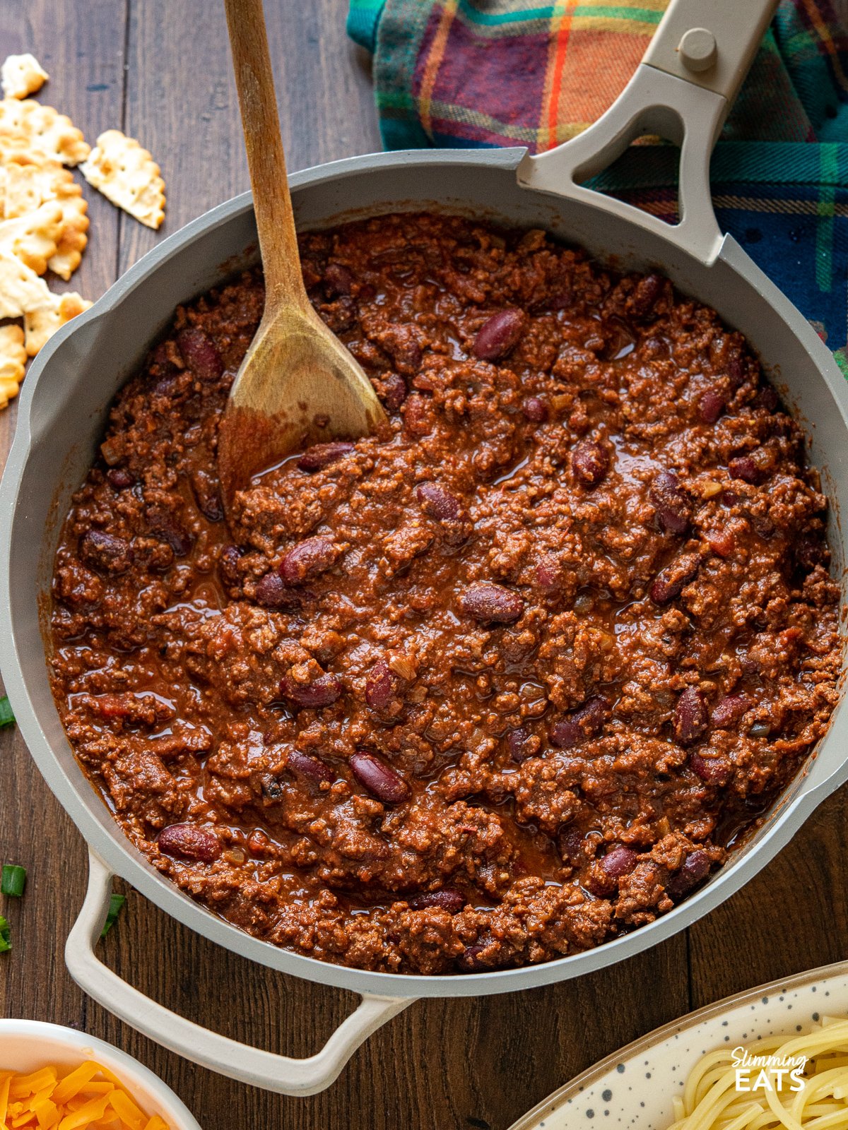 close up of Cincinnati Chili in frying pan with wooden spoon, sides and toppings place around pan